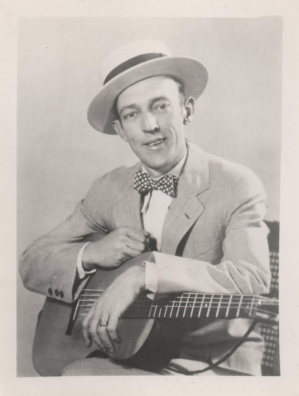 guitar player 桌布 壁紙 — 一位戴著軟呢帽和波點領結的微笑男子，在復古工作室肖像中抱著一把原聲吉他。