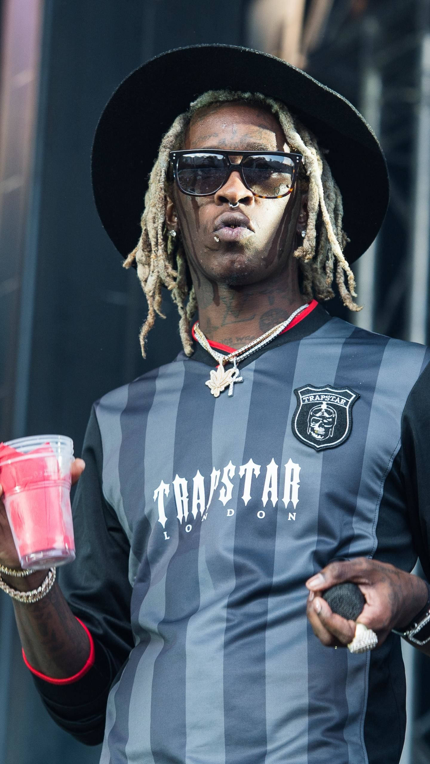 Thug 桌布 壁紙 — 一位戴著辮子頭和太陽眼鏡的饒舌歌手，穿著條紋 'Trapstar London' 襯衫，手持粉紅色飲料和麥克風。