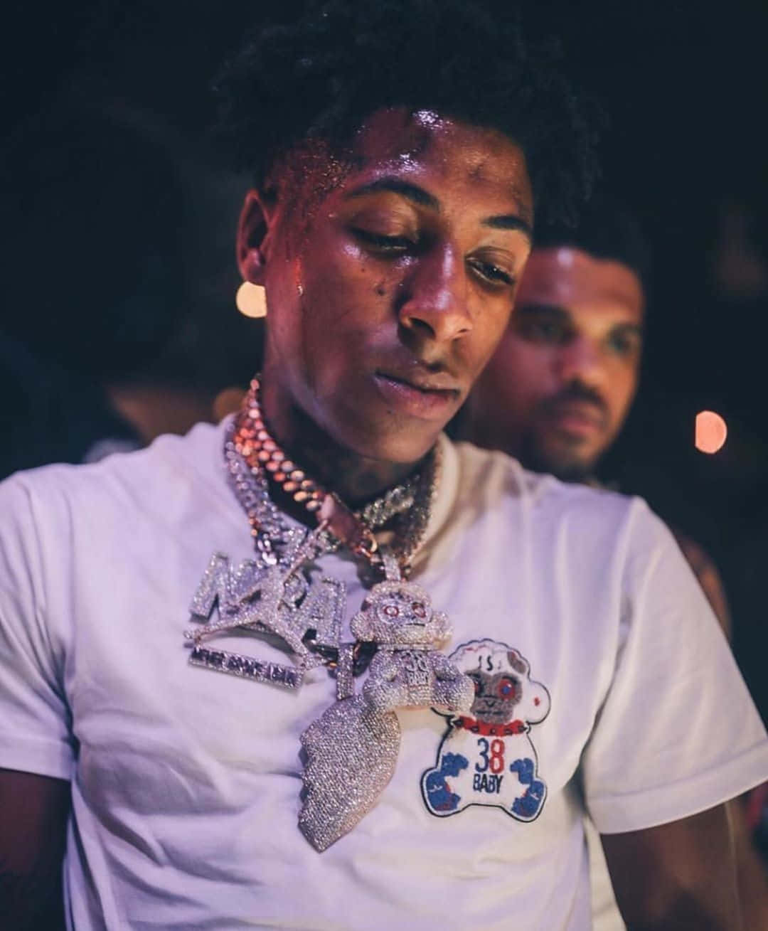 Youngboy 桌布 壁紙 — 一位戴著閃亮珠寶、身穿白色圖案T恤的年輕男子，在燈光昏暗、帶有散景燈光的俱樂部裡低頭看著。