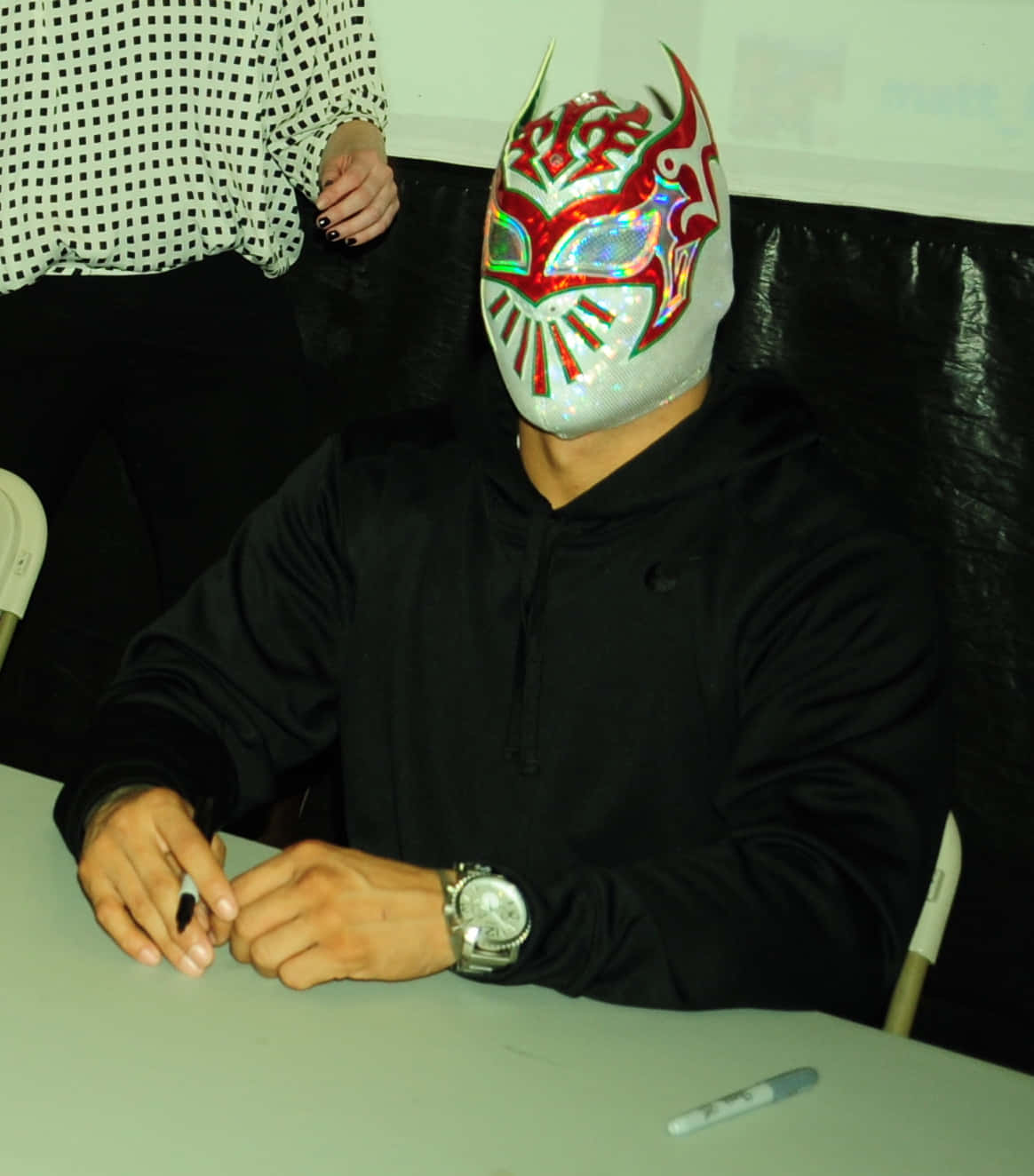 WWE 桌布 壁紙 — 一位戴著閃亮紅、綠、銀色面具的蒙面摔角手坐在室內桌子旁簽名。