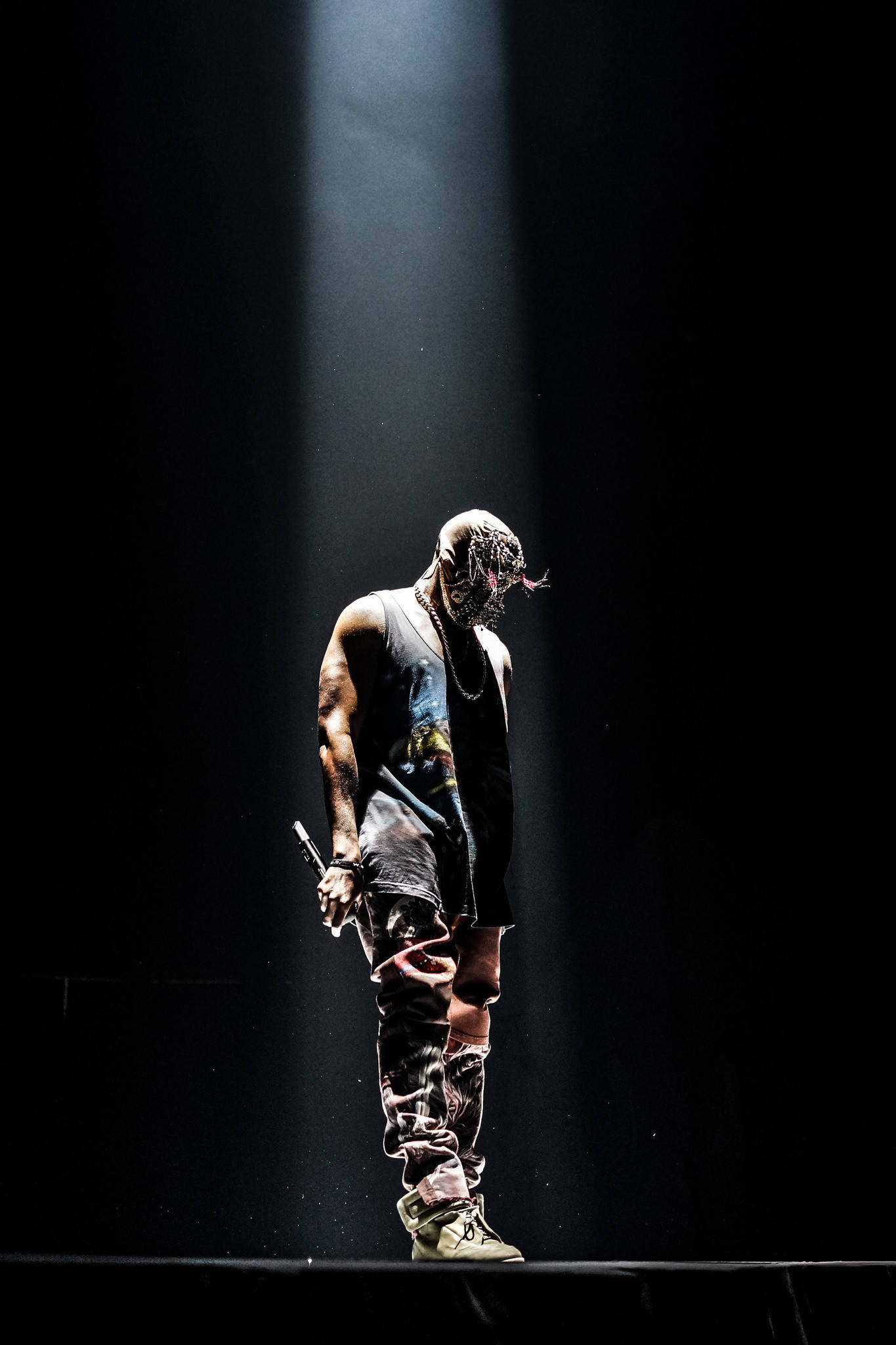 Kanye 桌布 壁紙 — 一位戴著面具、穿著破爛服裝的表演者獨自站在黑暗舞台上的單一聚光燈下。