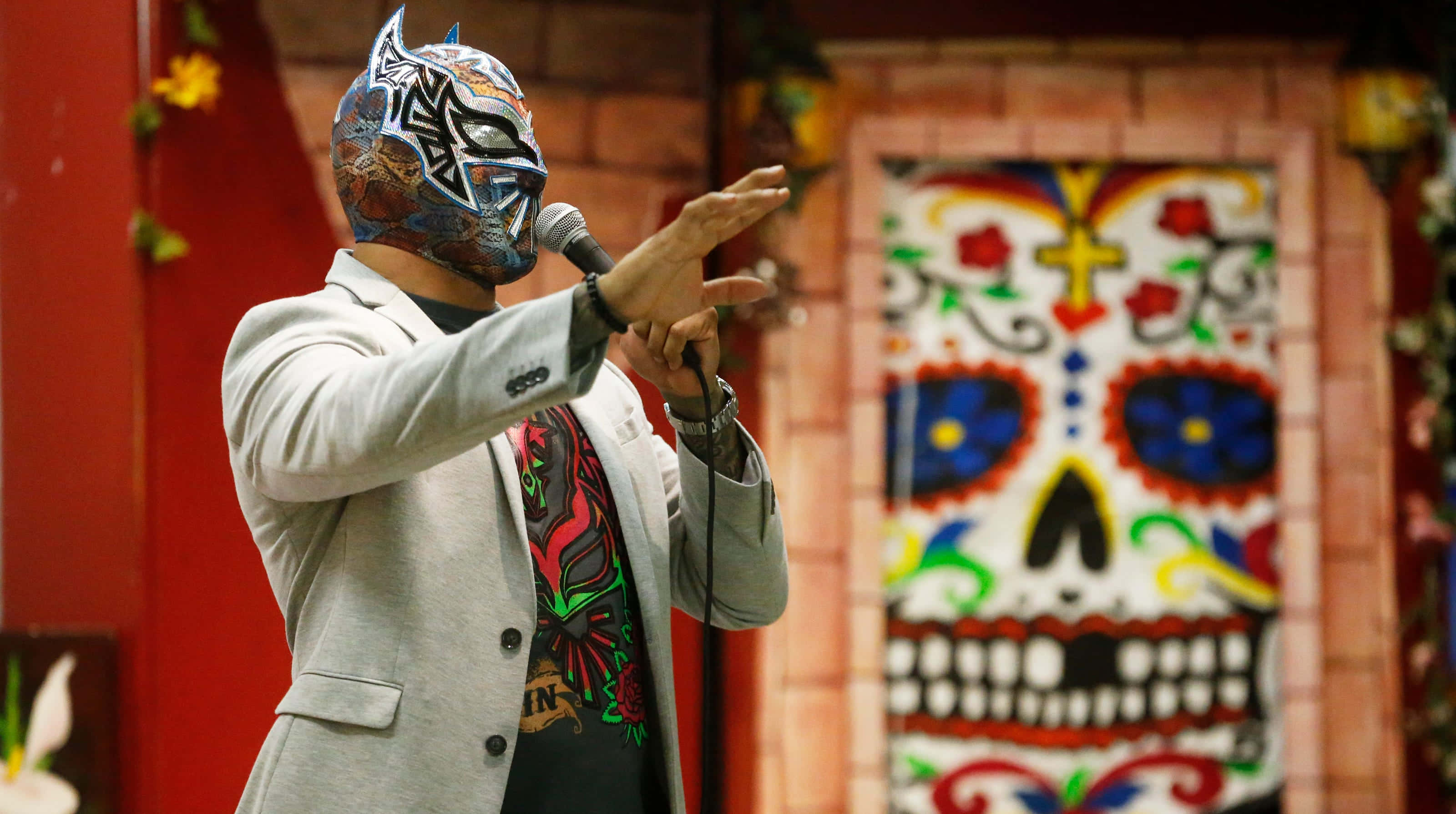 WWE 桌布 壁紙 — 一位戴著面具、身穿灰色西裝的摔角手，在色彩繽紛的糖骷髏背景下，手持麥克風做出戲劇性的手勢。