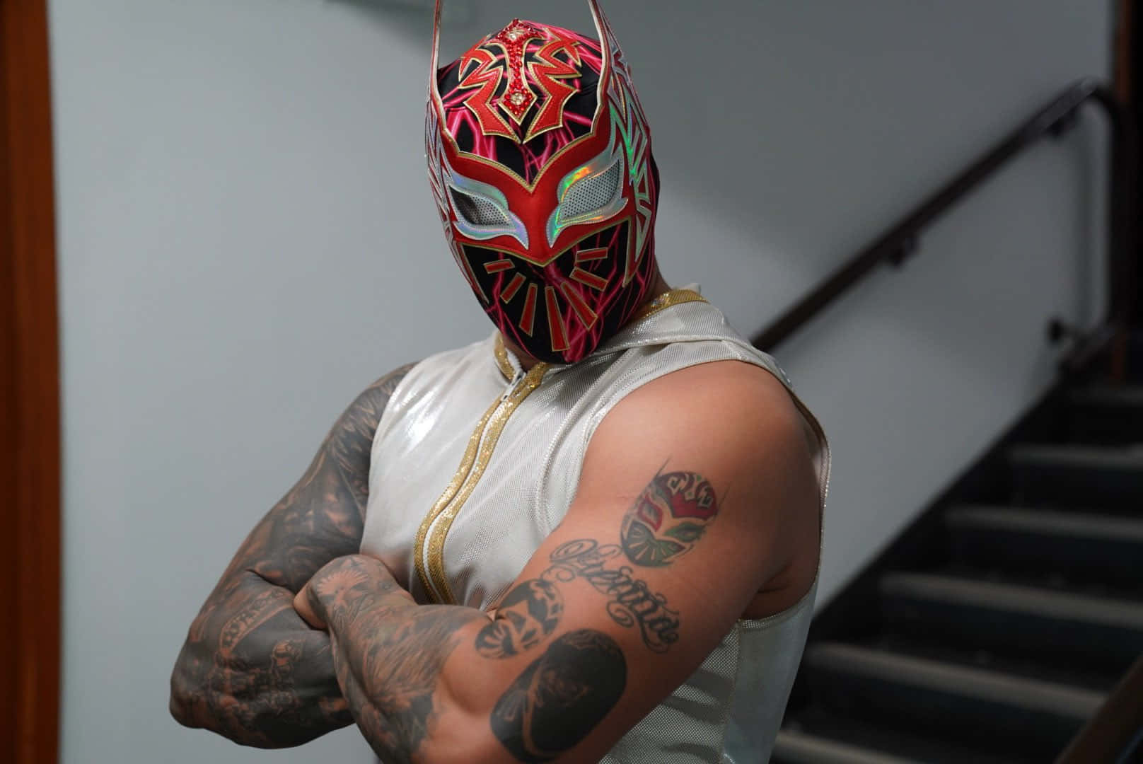 WWE 桌布 壁紙 — 一位戴著面具的摔角手，臉上繪有精細的紅黑圖案，自信地交叉雙臂站在樓梯間。