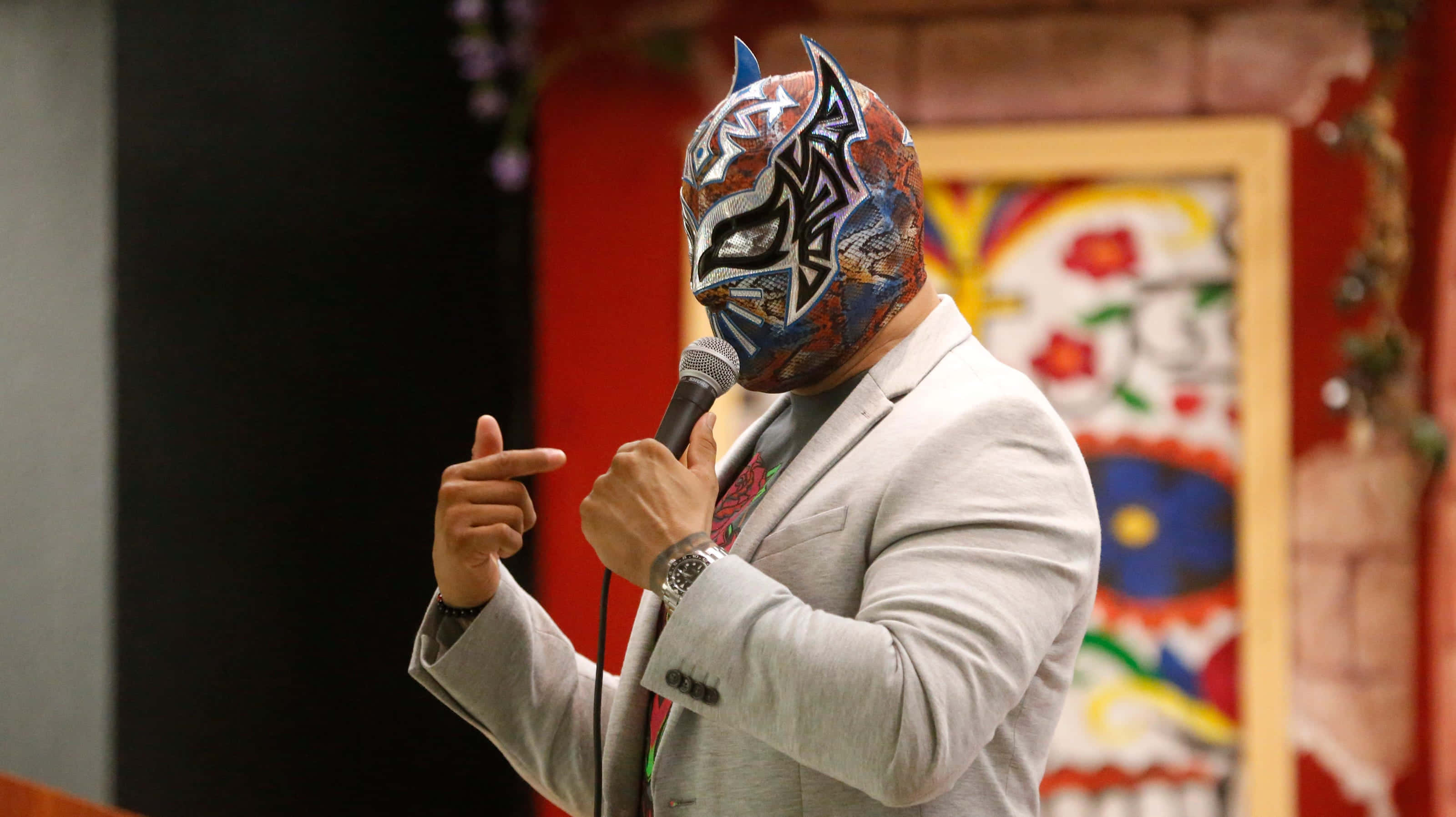 WWE 桌布 壁紙 — 一位戴著面具的摔角手身穿筆挺西裝，手持麥克風，在色彩繽紛的背景前以戲劇性的強度指向某處。