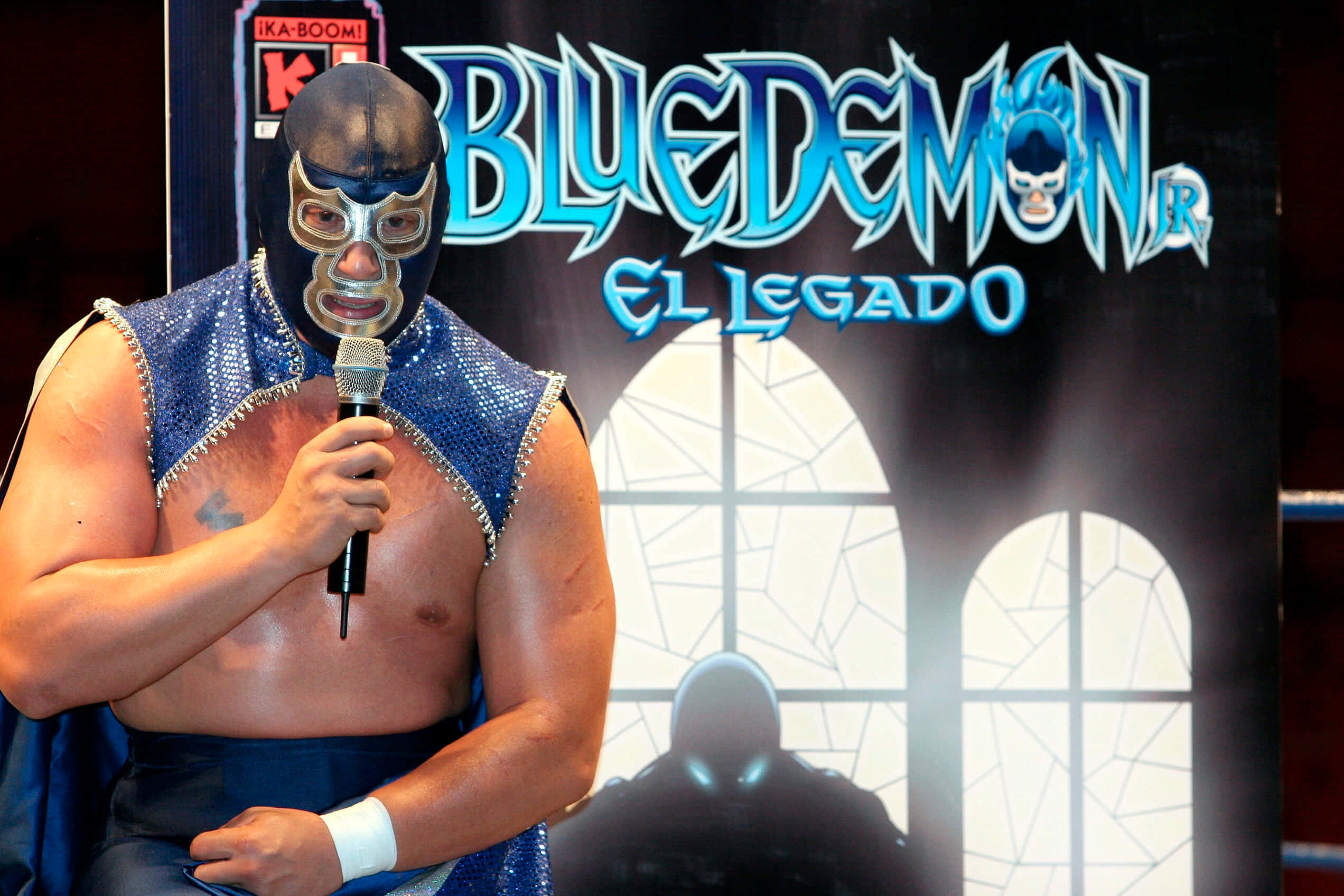 Mexican 桌布 壁紙 — 一位戴著面具的摔角手身穿藍色亮片服裝，在『Blue Demon El Legado』背景前手持麥克風。