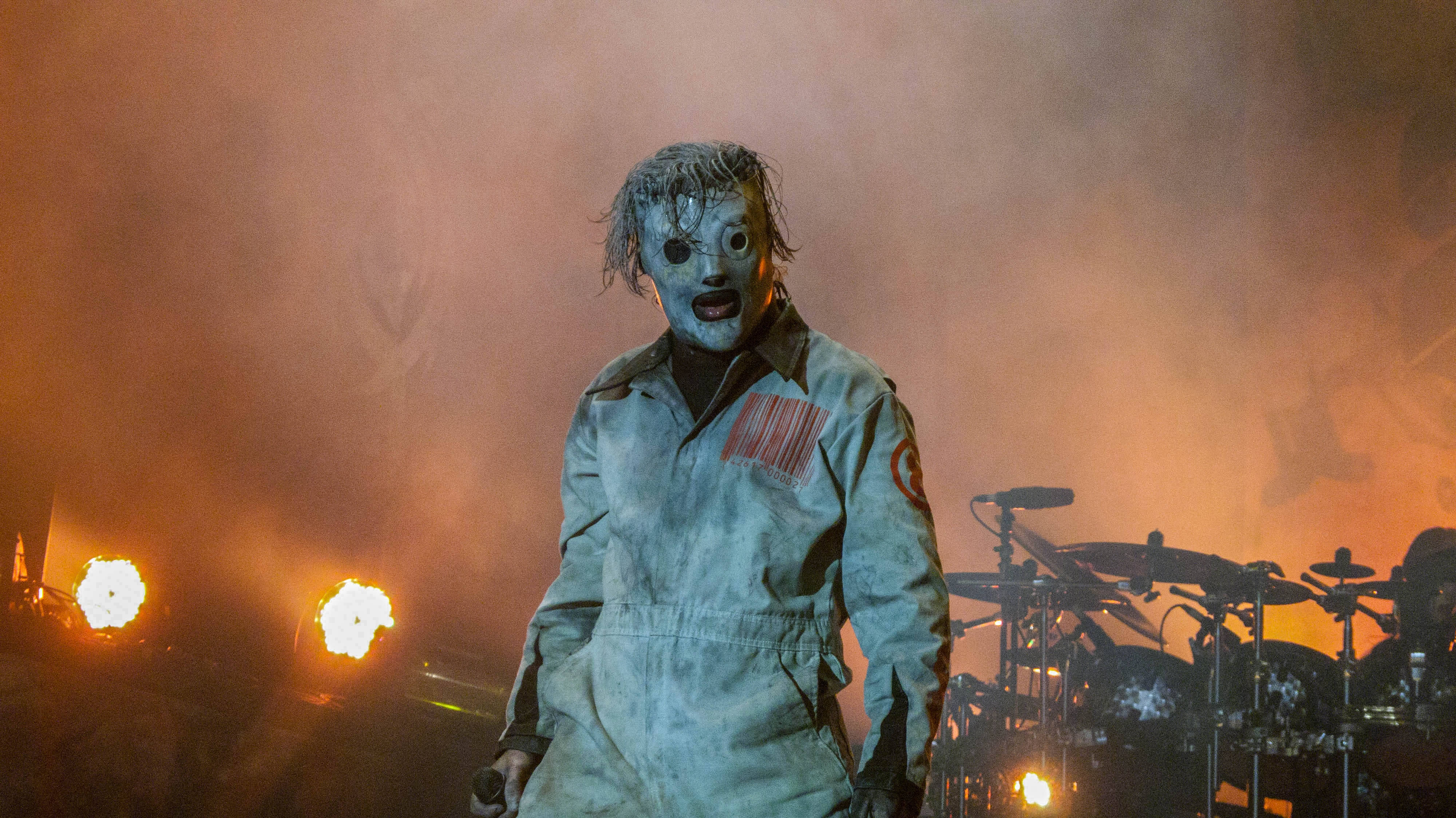 Slipknot 桌布 壁紙 — 一位戴著面具的表演者穿著骯髒的連身衣，在煙霧彌漫的舞台上，沐浴在火熱的橙色燈光中，熱情地歌唱。