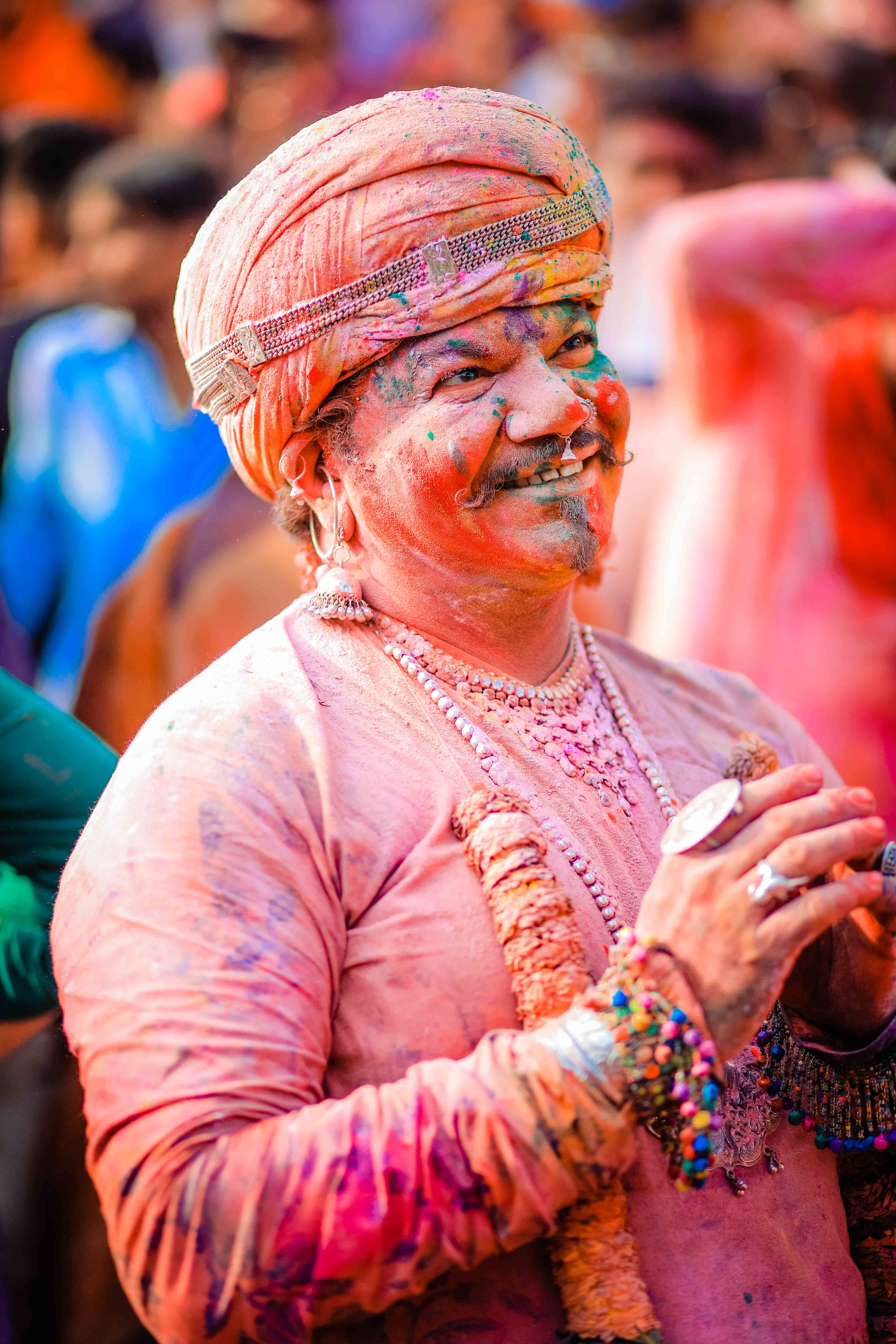 Holi 桌布 壁紙 — 一位戴著頭巾和串珠首飾的快樂男子，在節日慶祝中，彩色粉末覆蓋他的臉和衣服，他露出燦爛的笑容。