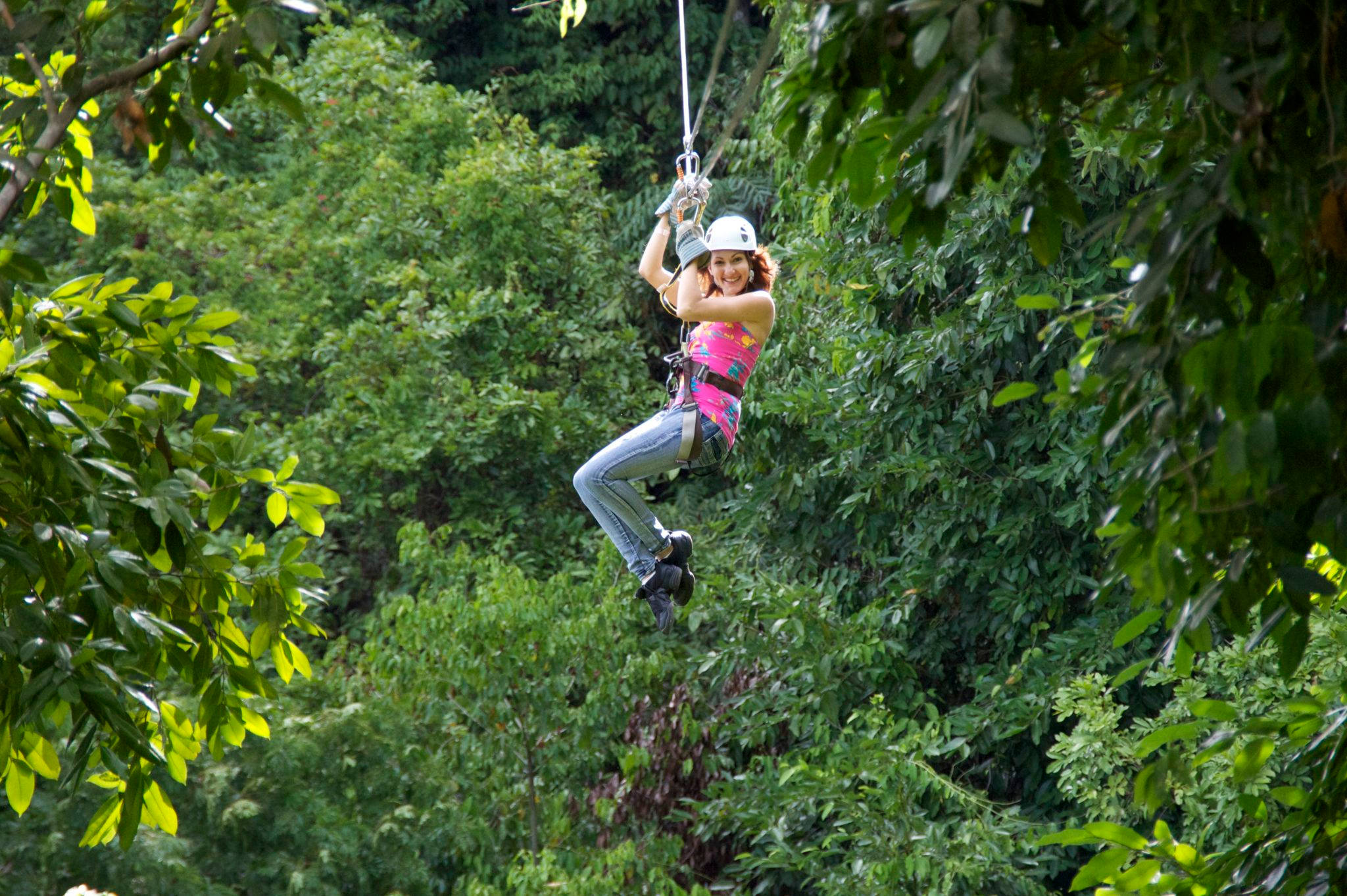 牙買加 桌布 壁紙 — 一位戴著頭盔微笑的女性在陽光明媚的日子裡，沿著翠綠的森林樹冠滑索道。
