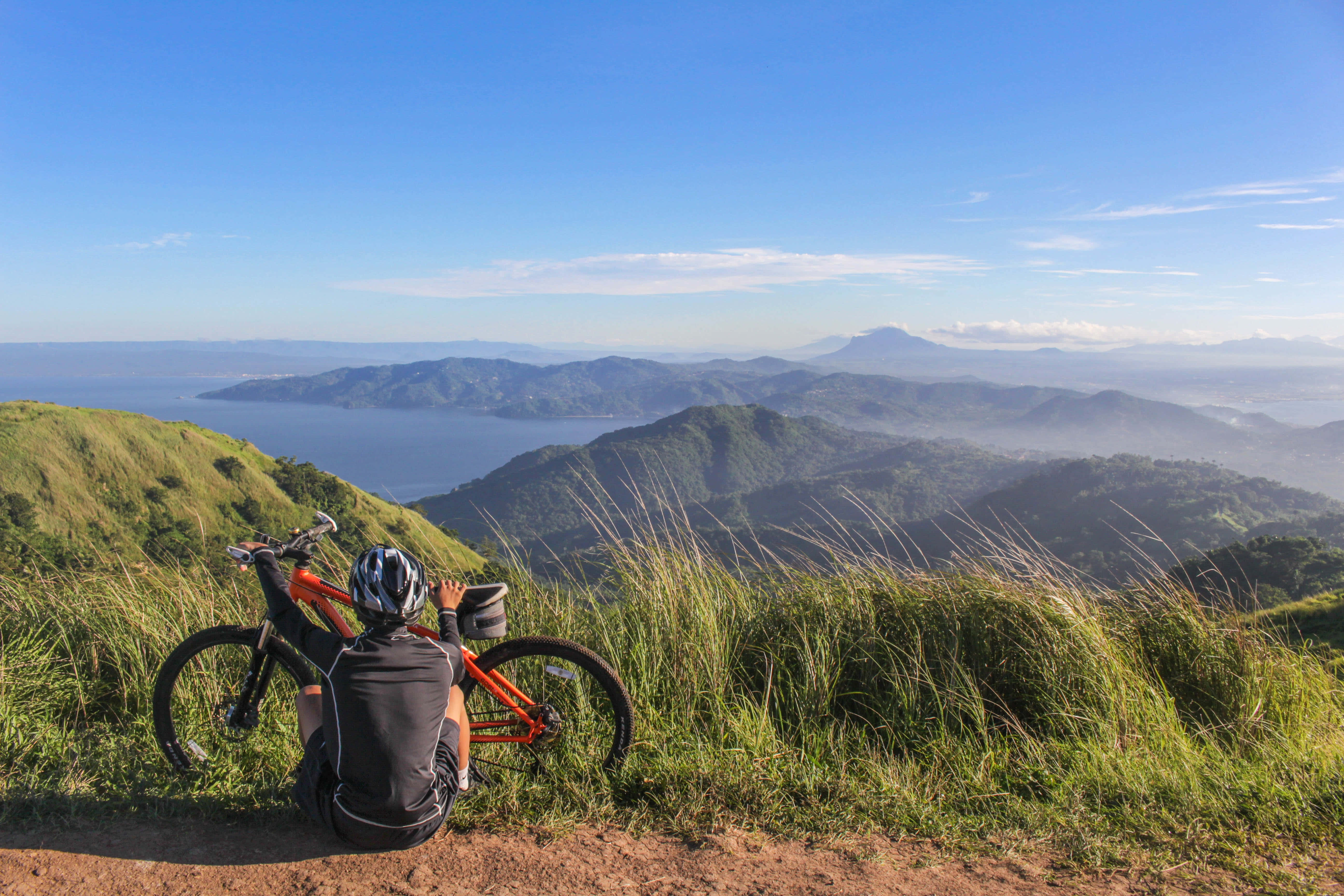Outdoor Activities 桌布 壁紙 — 一位戴著頭盔的自行車手在長滿草的山頂上，在他們的橙色自行車旁邊休息，俯瞰著廣闊的海洋和層層疊疊的山脈，天空清澈湛藍。