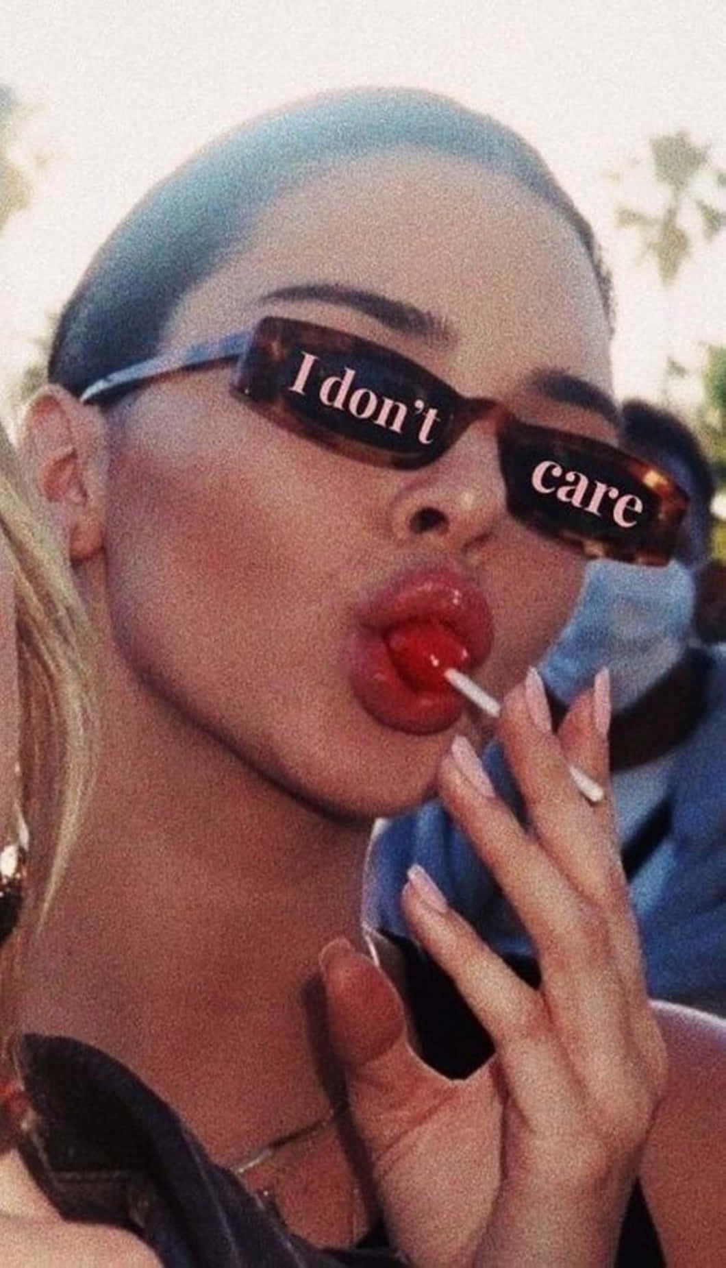 Girl Aesthetic 桌布 壁紙 — 一位戴著顯示「I don't care」太陽眼鏡的女性，在戶外擺姿勢咬著紅色棒棒糖。