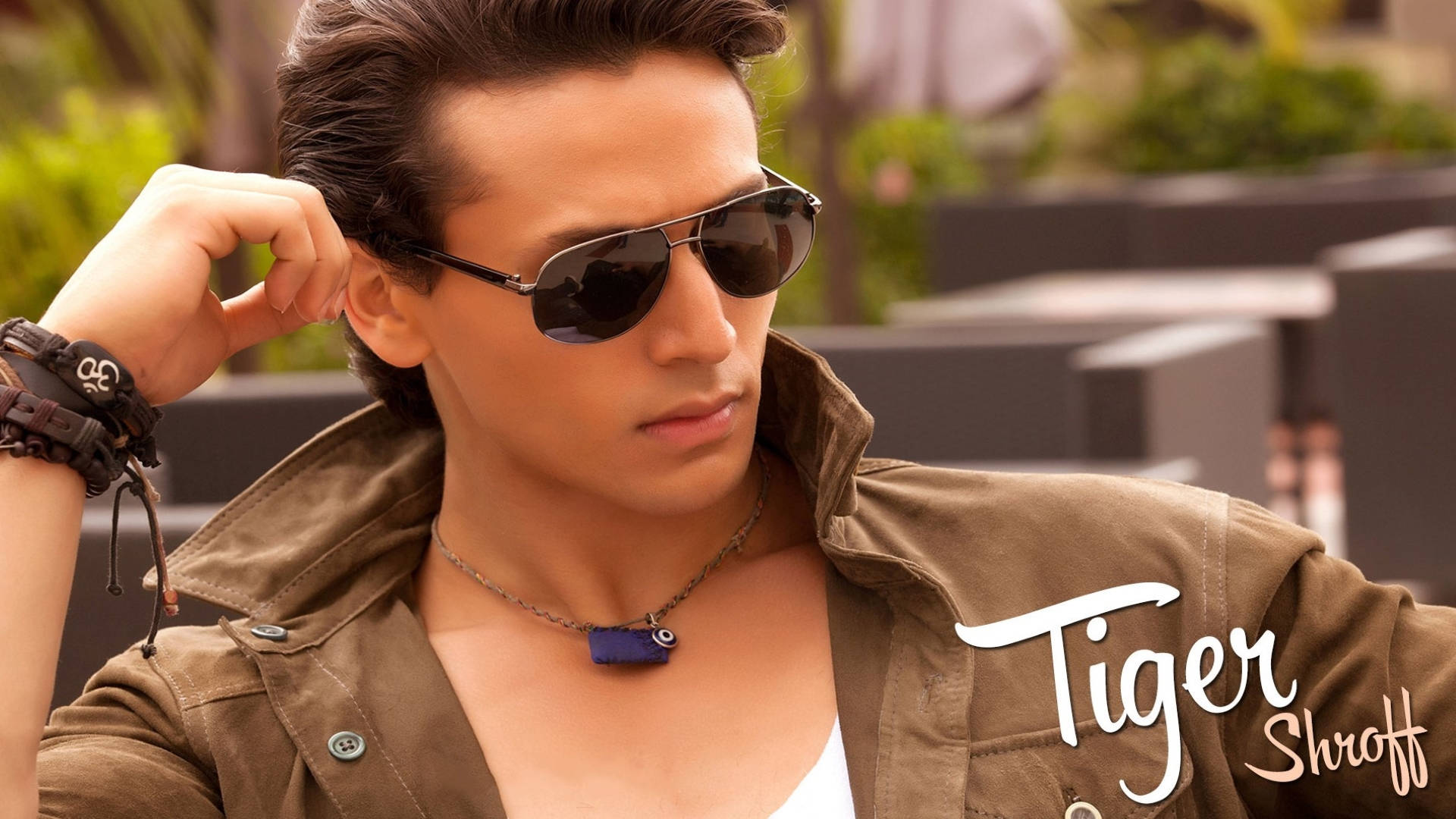 Tiger Shroff 桌布 壁紙 — 一位戴著飛行員太陽眼鏡和棕色外套的時尚男士，在柔和的日光下自信地於戶外擺姿勢，周圍有綠植和現代家具。