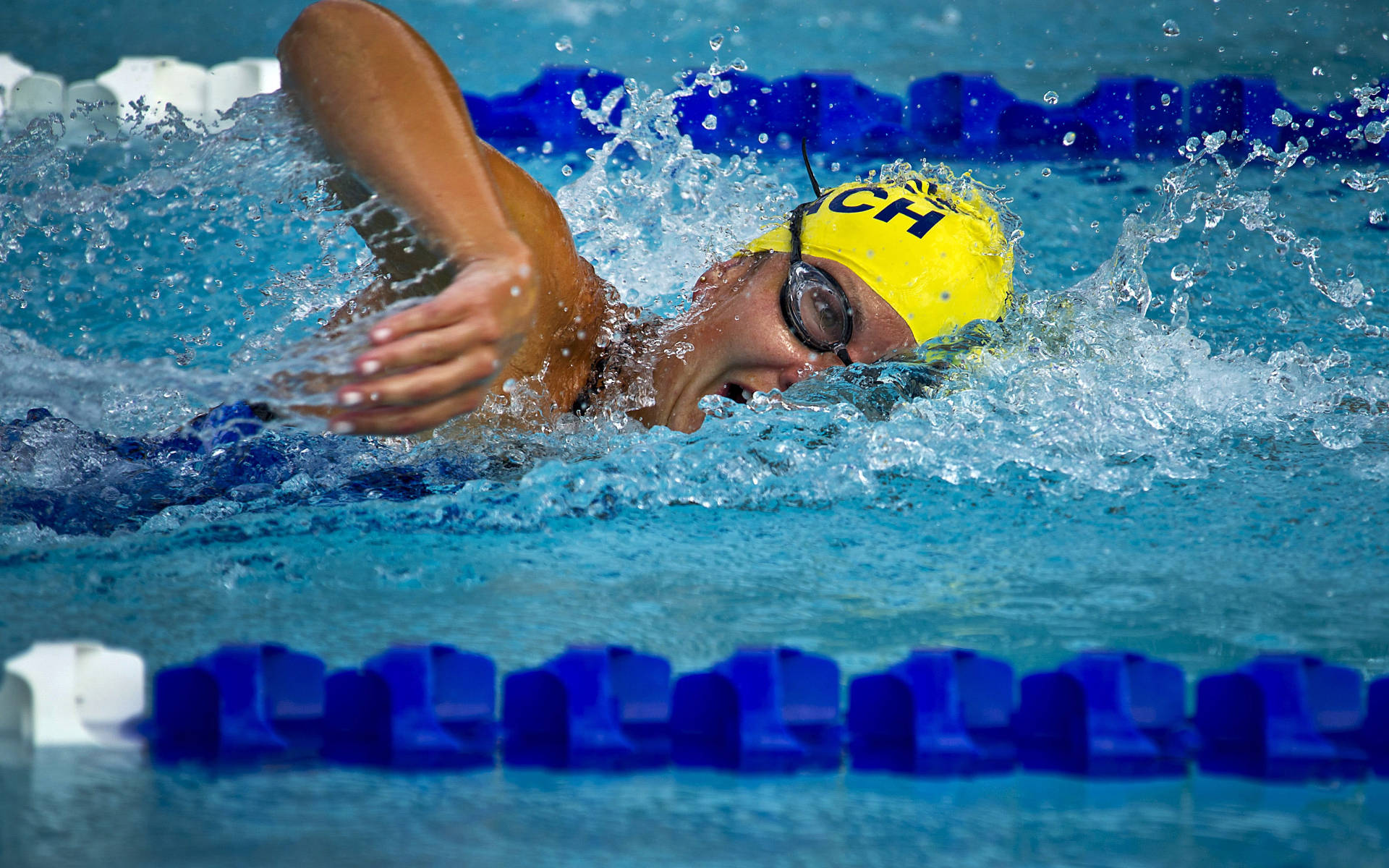 Swimming 桌布 壁紙 — 一位戴著黃色泳帽和泳鏡的游泳者，在充滿活力的藍色泳池水中強而有力地執行自由式划水。
