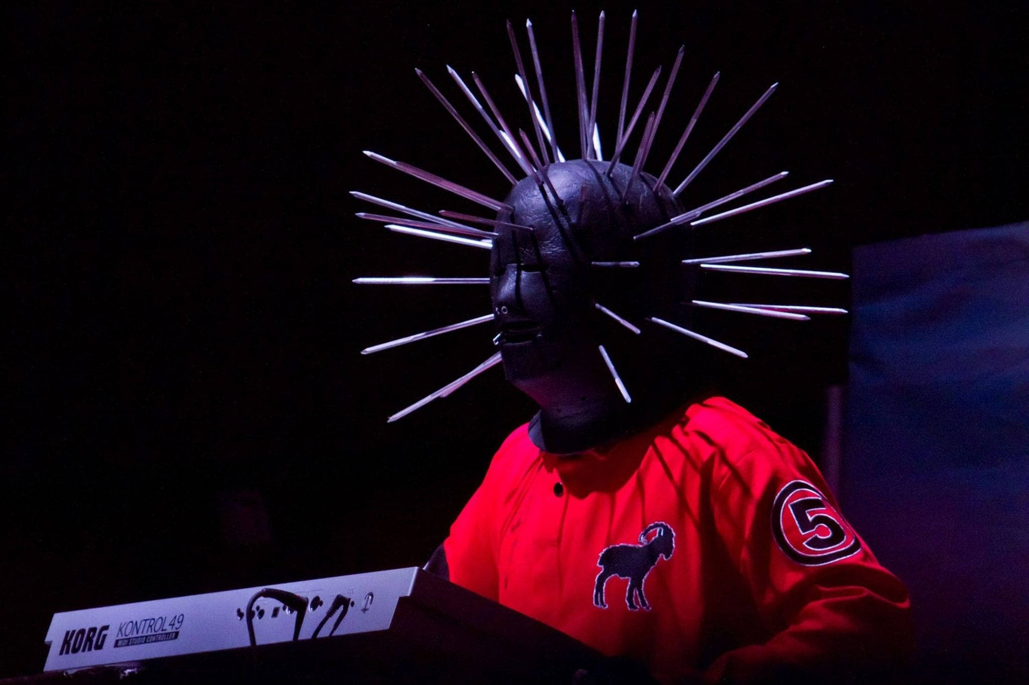craig jones 桌布 壁紙 — 一位戴著黑色尖刺面具、身穿紅色外套的音樂家在戲劇性的舞台燈光下演奏Korg鍵盤。