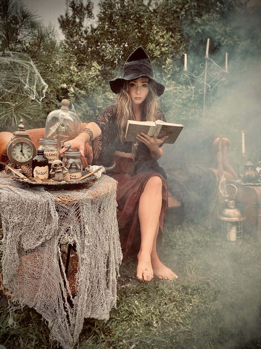 Witch Costumes 桌布 壁紙 — 一位戴著黑色帽子的女巫在迷霧花園中閱讀一本書，周圍環繞著神秘罐子、蠟燭和煙霧。