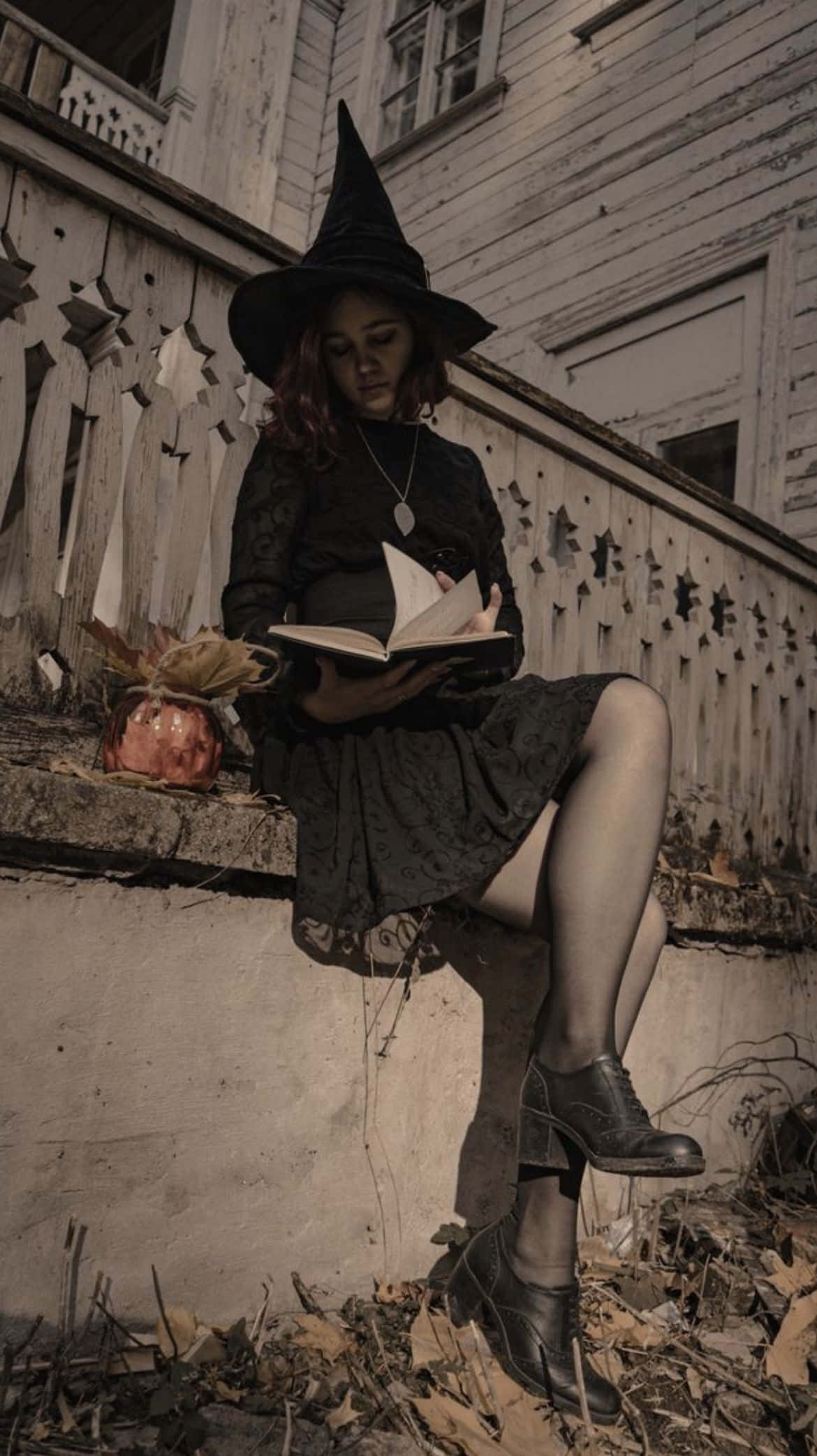 Witch Costumes 桌布 壁紙 — 一位戴著黑色帽子的女巫坐在佈滿秋葉的風化門廊上閱讀一本書。