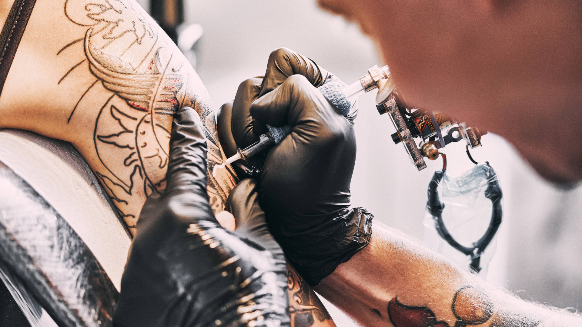 Tattoo 桌布 壁紙 — 一位戴著黑色手套的紋身藝術家，正使用專業紋身機，仔細地將墨水注入客戶手臂上精緻的龍形圖案。