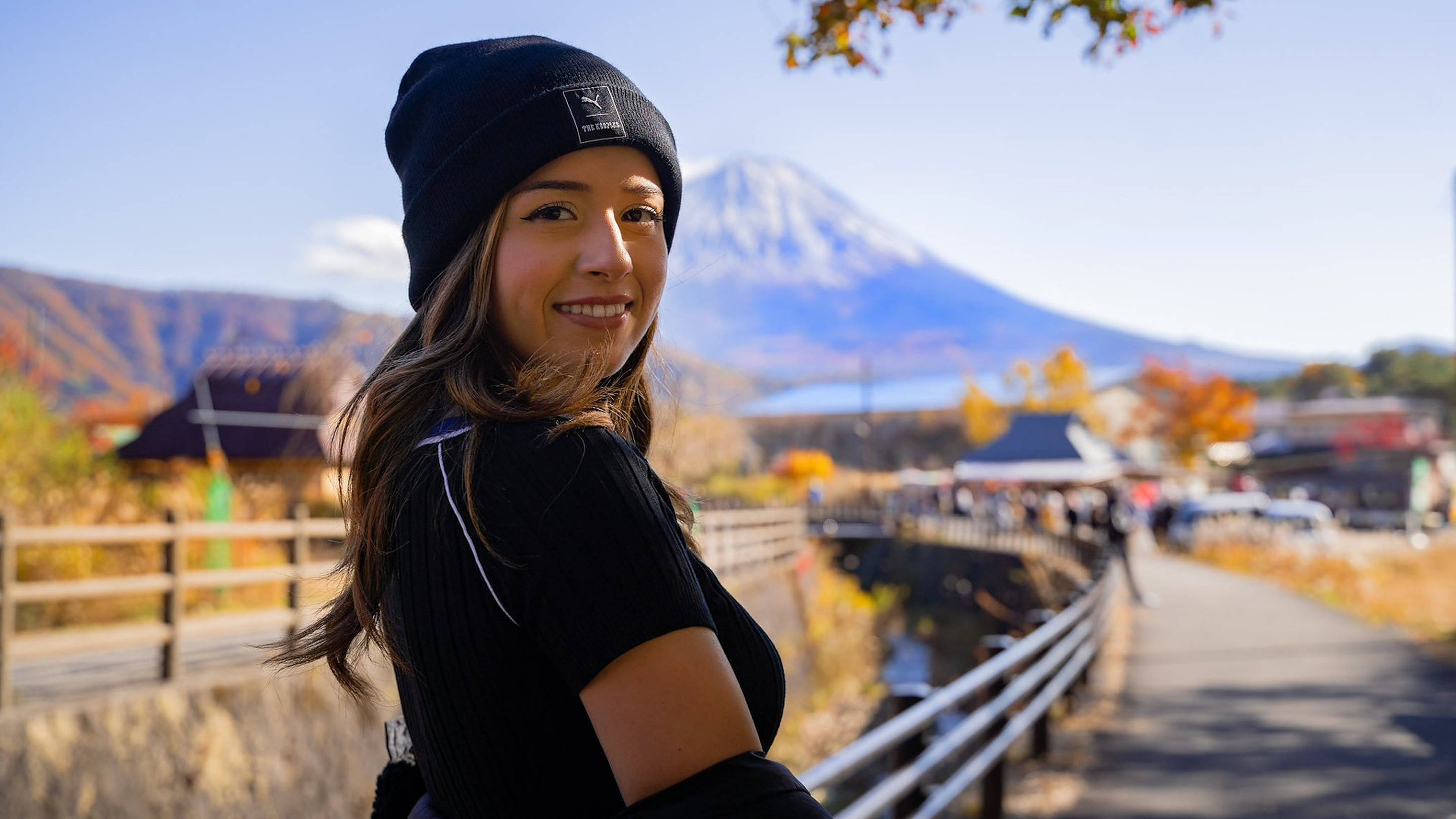 pokimane 桌布 壁紙 — 一位戴著黑色毛帽的微笑女士轉向鏡頭，背景是富士山和秋天的樹葉。