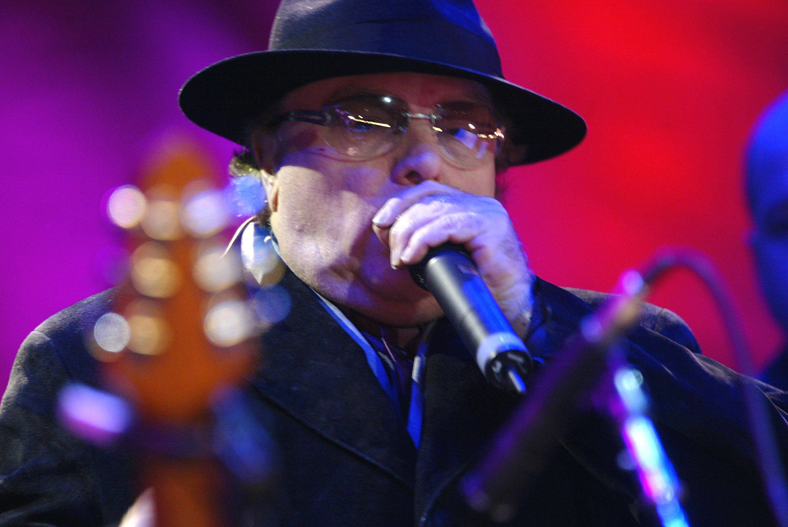 Van Morrison 桌布 壁紙 — 一位戴著黑色軟呢帽和眼鏡的音樂家在充滿活力的紫色和紅色舞台燈光下熱情地對著麥克風唱歌。