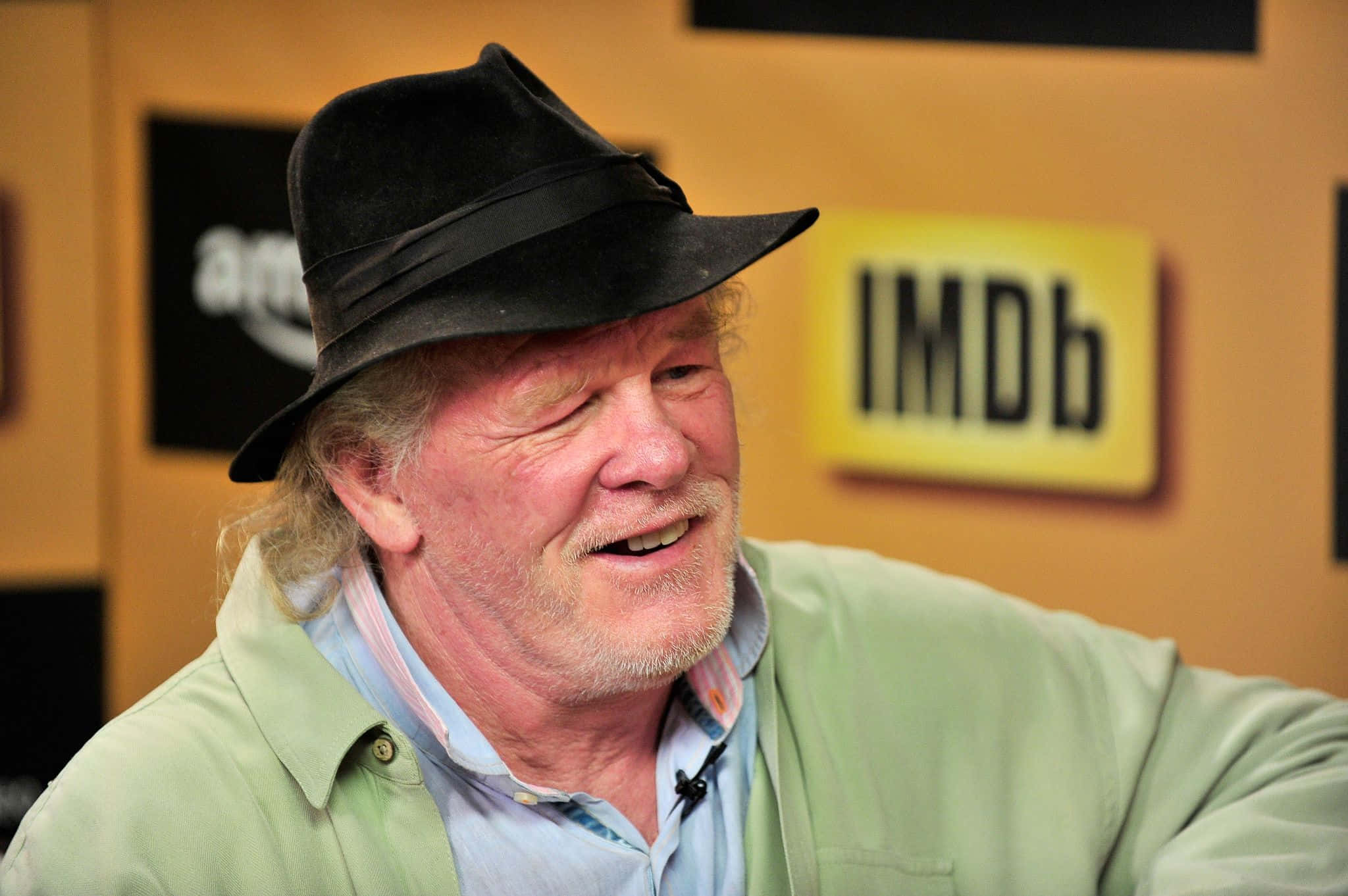 Nick Nolte 桌布 壁紙 — 一位戴著黑色軟呢帽和綠色外套的微笑年長男性在 IMDb 背景前溫暖地大笑。