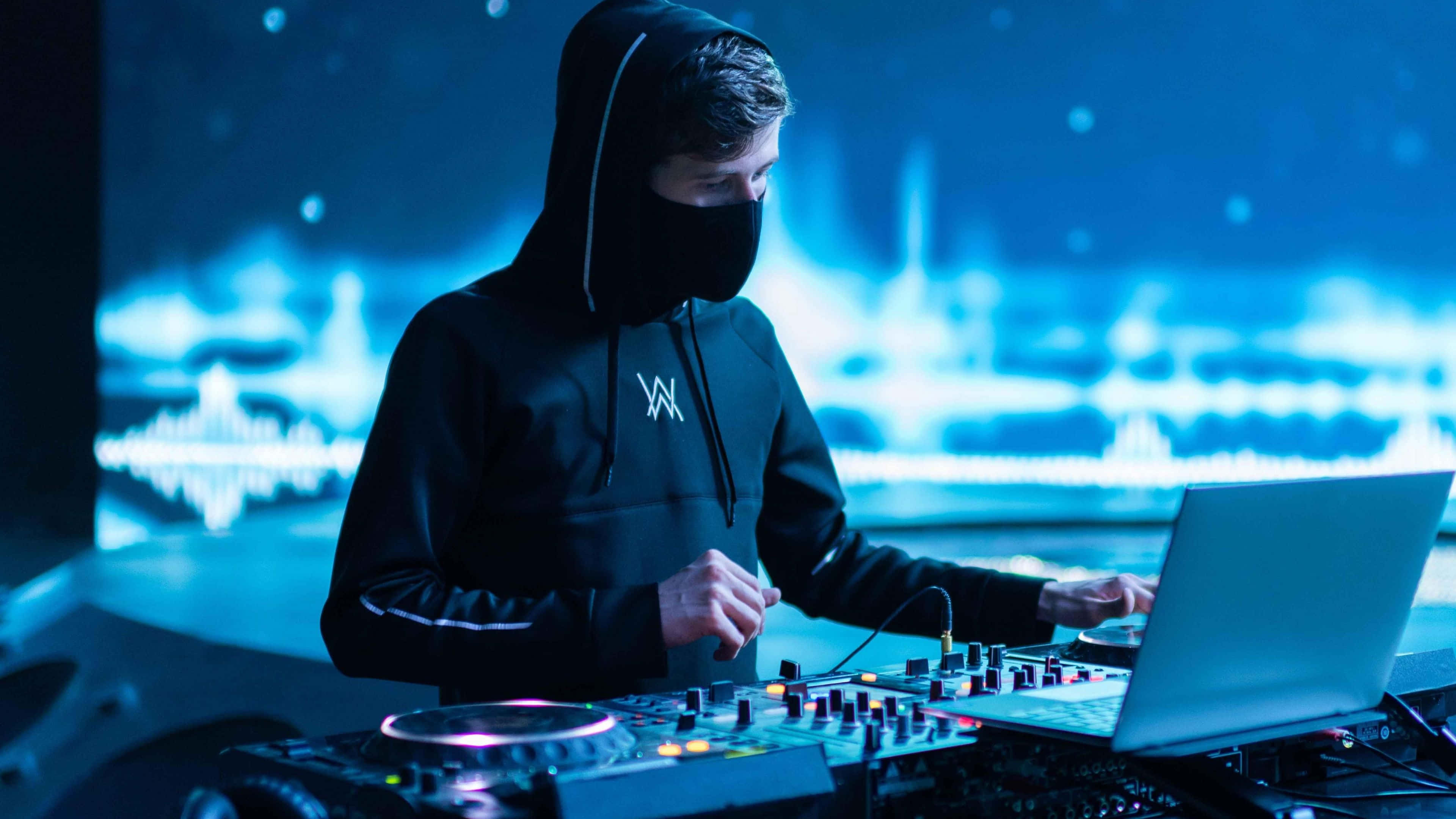 艾倫·沃克 桌布 壁紙 — 一位戴著黑色面具、身穿黑色連帽衫的DJ，在藍色霓虹燈下用發光的控制器混音。