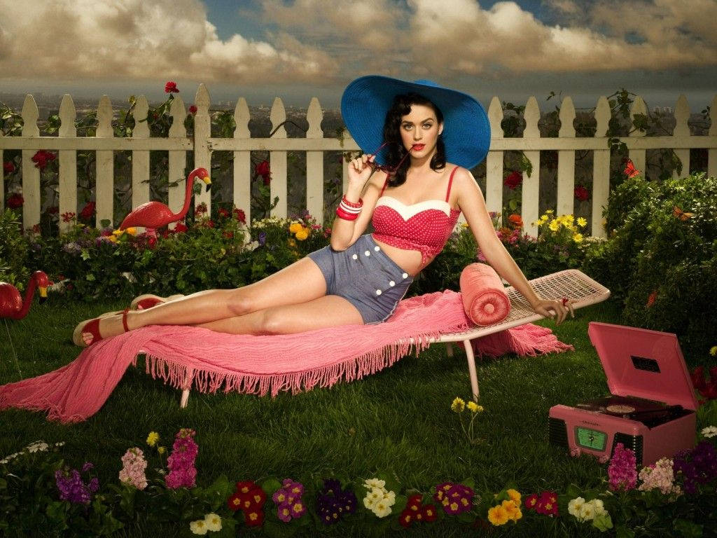 Perry 桌布 壁紙 — 一位戴藍色帽子、穿紅色上衣的女性在一個充滿活力的花園裡，躺在粉紅色毯子上休息，花園有白色柵欄和粉紅色火烈鳥。
