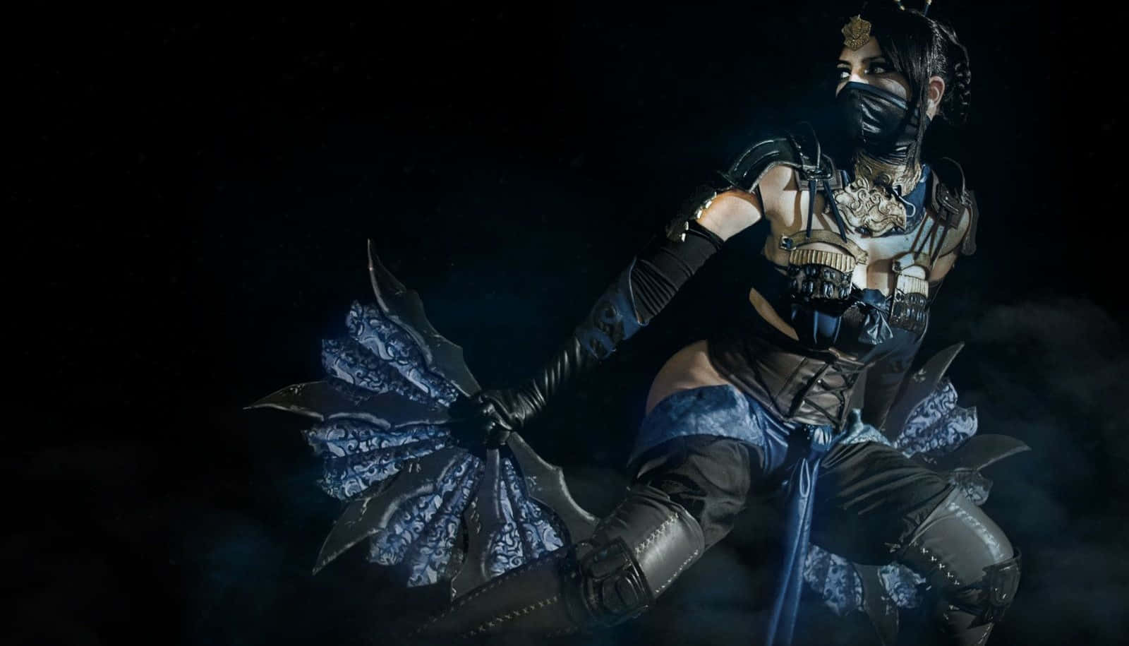 Mortal Kombat Kitana 桌布 壁紙 — 一位戴面具的戰士身穿黑色盔甲，在煙霧繚繞的黑色背景下揮舞著帶刺的扇子，燈光效果戲劇化。