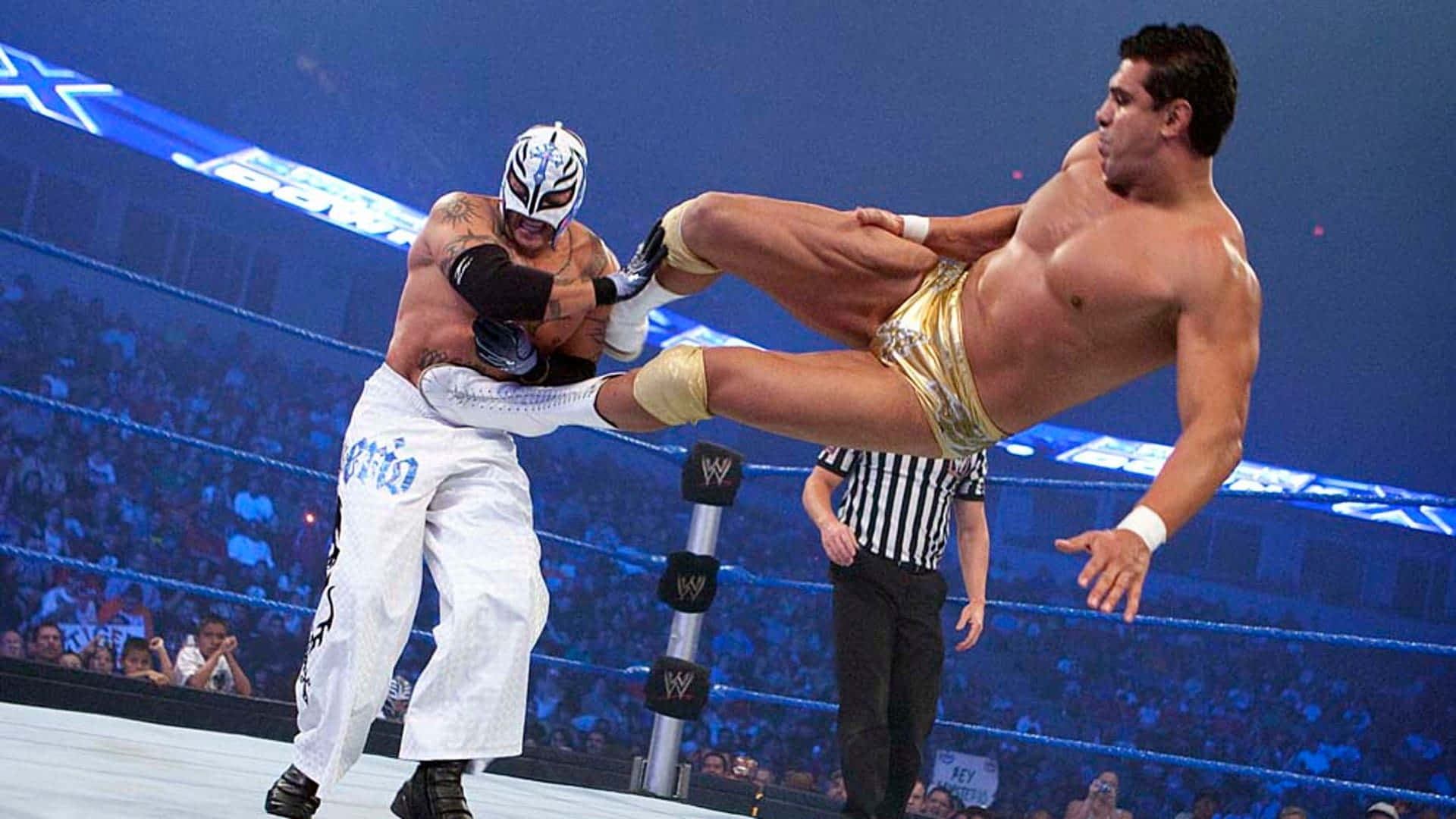 WWE 桌布 壁紙 — 一位戴面具的摔角手穿著白色褲子，在明亮的場館燈光下於WWE比賽中對一位穿著金色短褲的對手使出高踢腿。