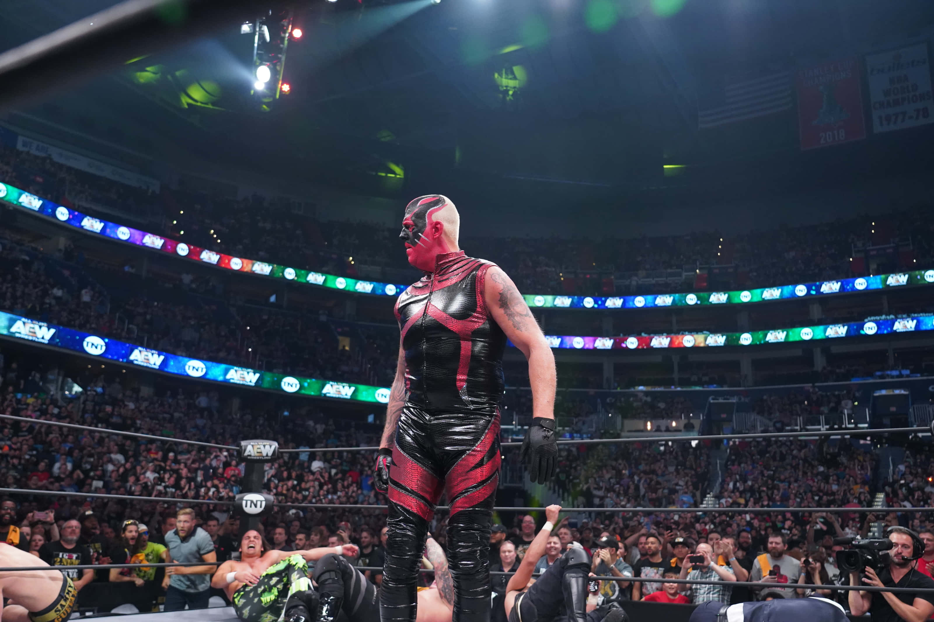 WWE 桌布 壁紙 — 一位戴面具的摔角手身穿紅黑戰衣，在明亮的場館燈光下站在擂台上，台下觀眾歡呼雷動。