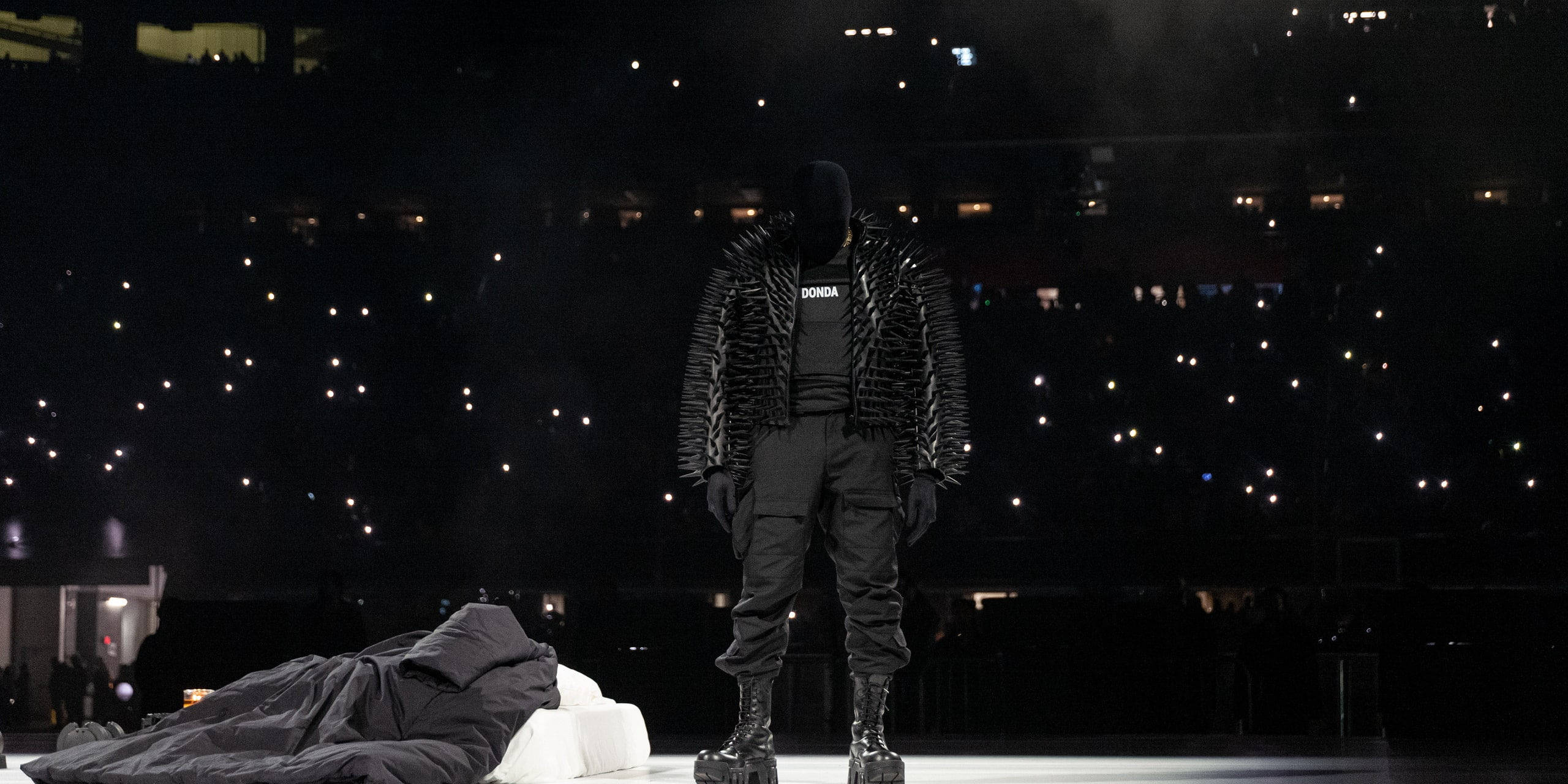 Kanye West Ye 桌布 壁紙 — 一位戴面具的表演者身穿帶有尖刺的黑色外套，獨自站在燈光昏暗的舞台上，身後黑暗的場館裡擠滿了舉著發光手機的人群。