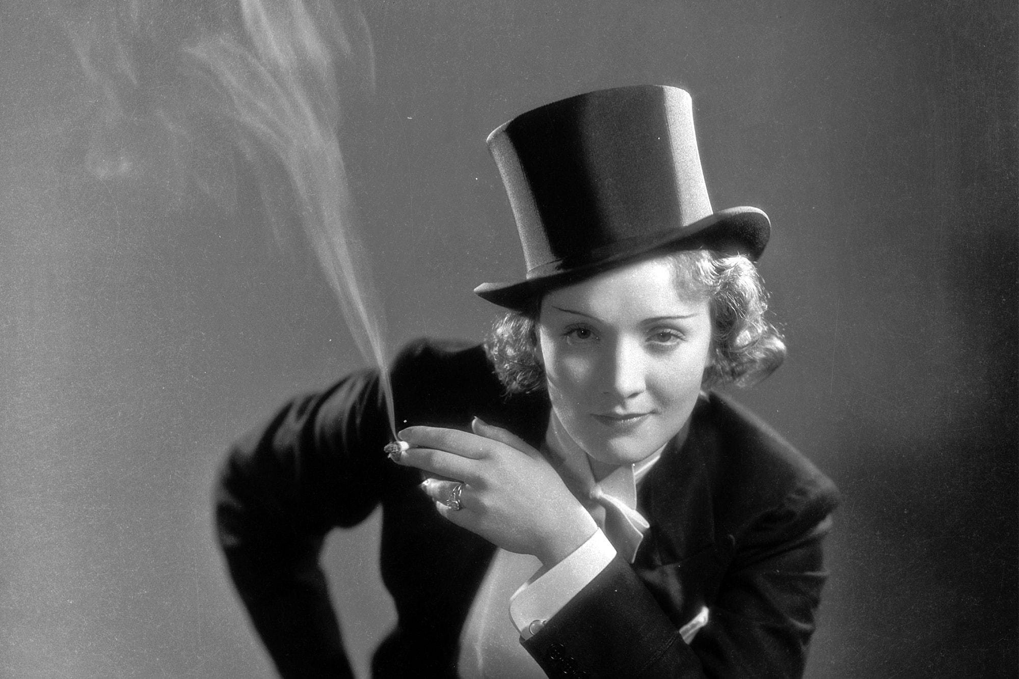 Marlene Dietrich 桌布 壁紙 — 一位戴高帽穿西裝的迷人女性，在柔和的灰色背景下，以自信的眼神抽煙。