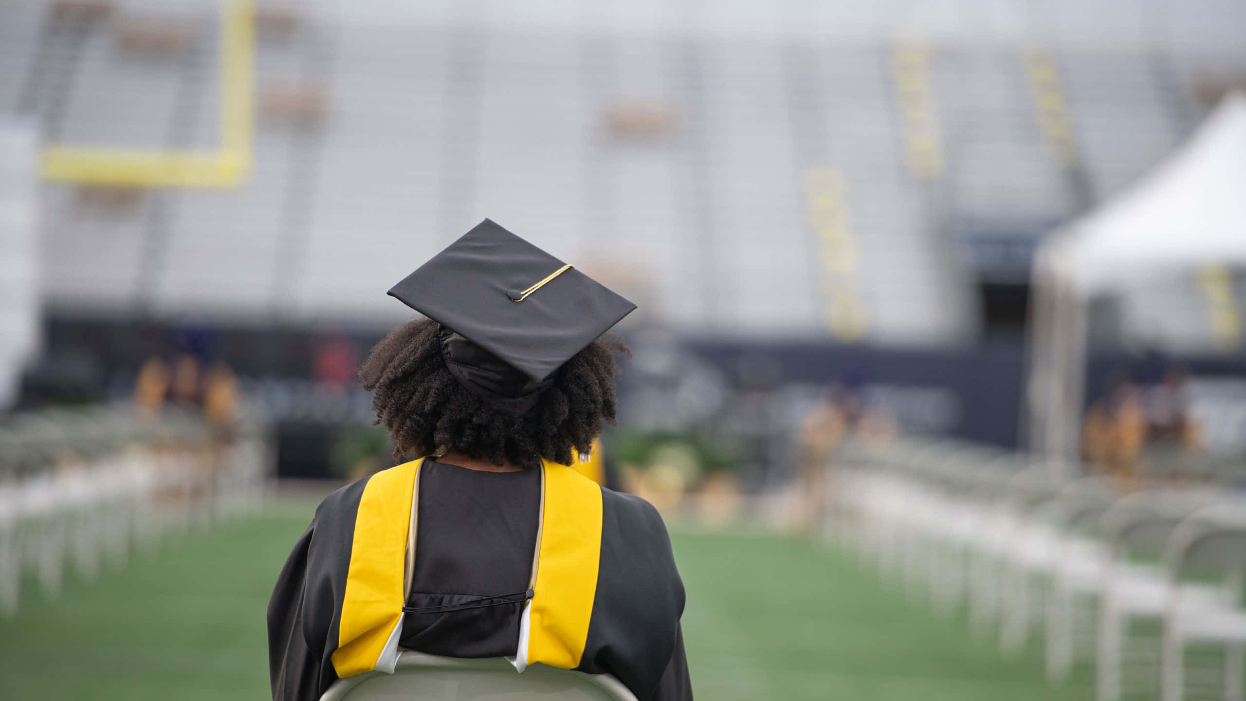 Graduation Ceremony 桌布 壁紙 — 一位戴黑色學士帽、披黃色肩帶的畢業生坐在椅子上，面向空無一人的運動場。