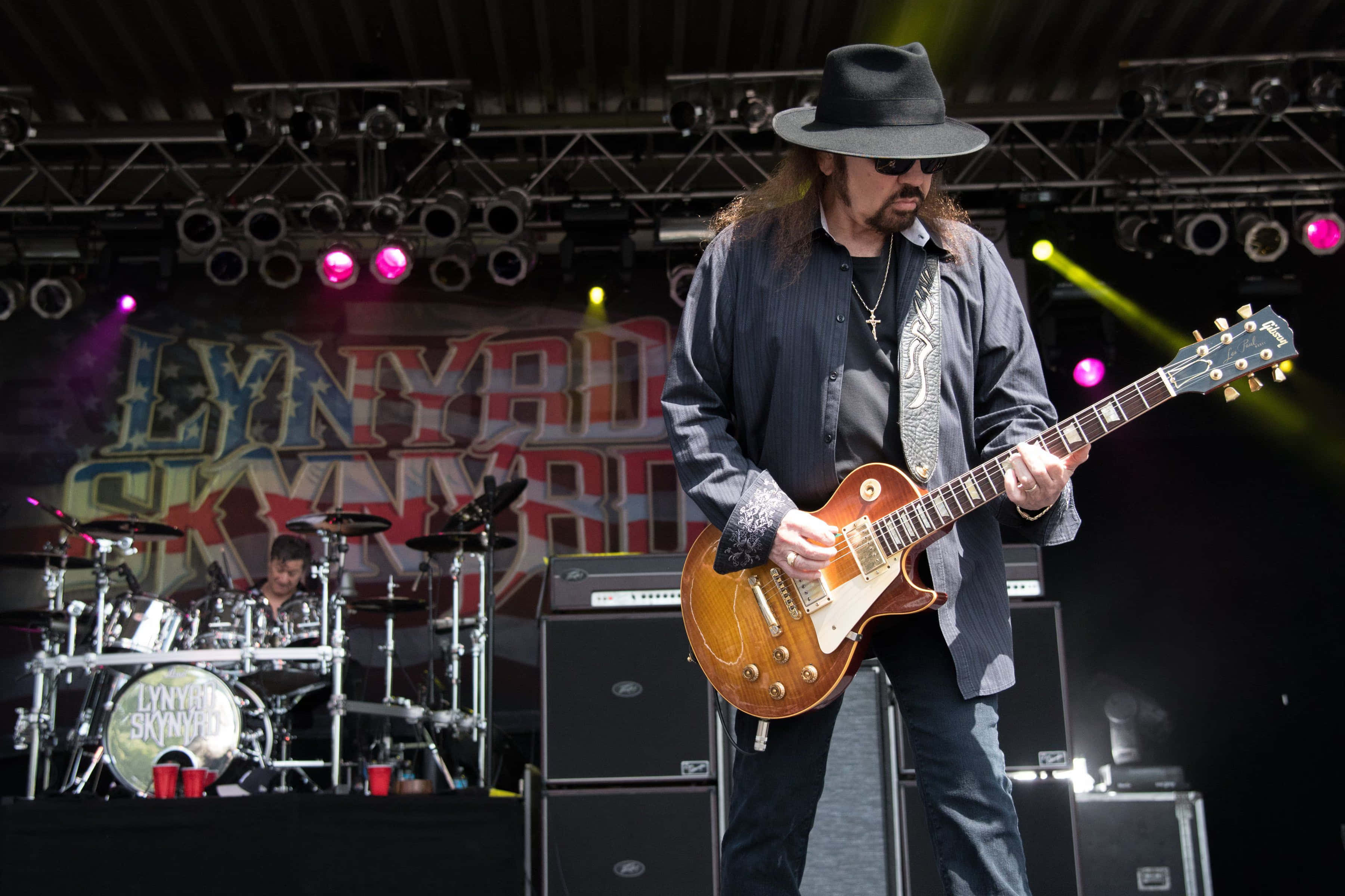 Gary Rossington 桌布 壁紙 — 一位戴黑色帽子和太陽眼鏡的吉他手在音樂會舞台上彈奏一把 sunburst 電吉他，身後有鼓手和色彩繽紛的舞台燈光。