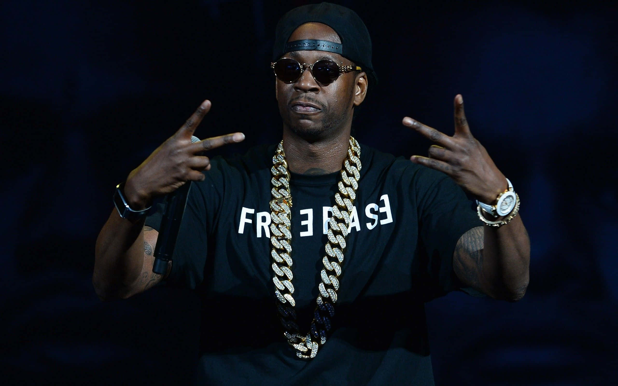 2 Chainz 桌布 壁紙 — 一位戴黑色帽子和太陽眼鏡的饒舌歌手，在舞台燈光下戴著金鍊和黑色T恤做手勢。