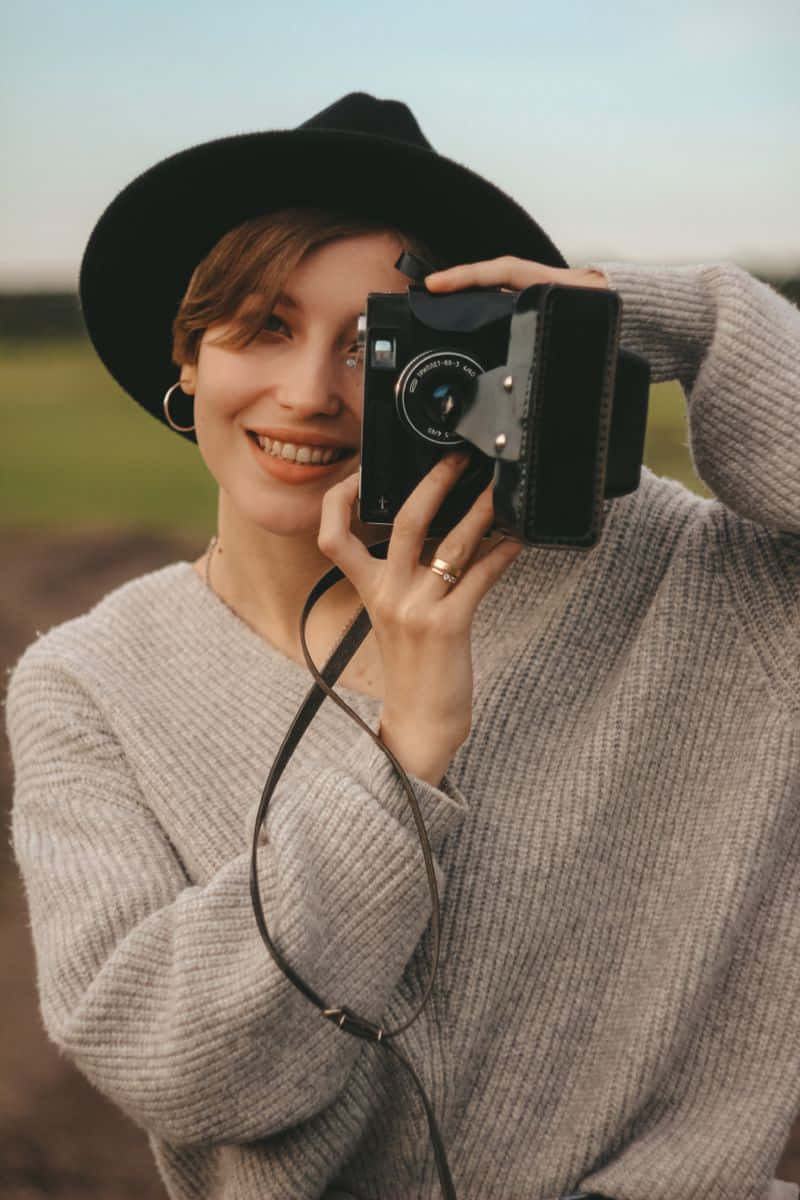 Holding Camera 桌布 壁紙 — 一位戴黑色帽子和灰色毛衣的微笑女士，在黃金時刻站在草地上，手持一部復古相機。