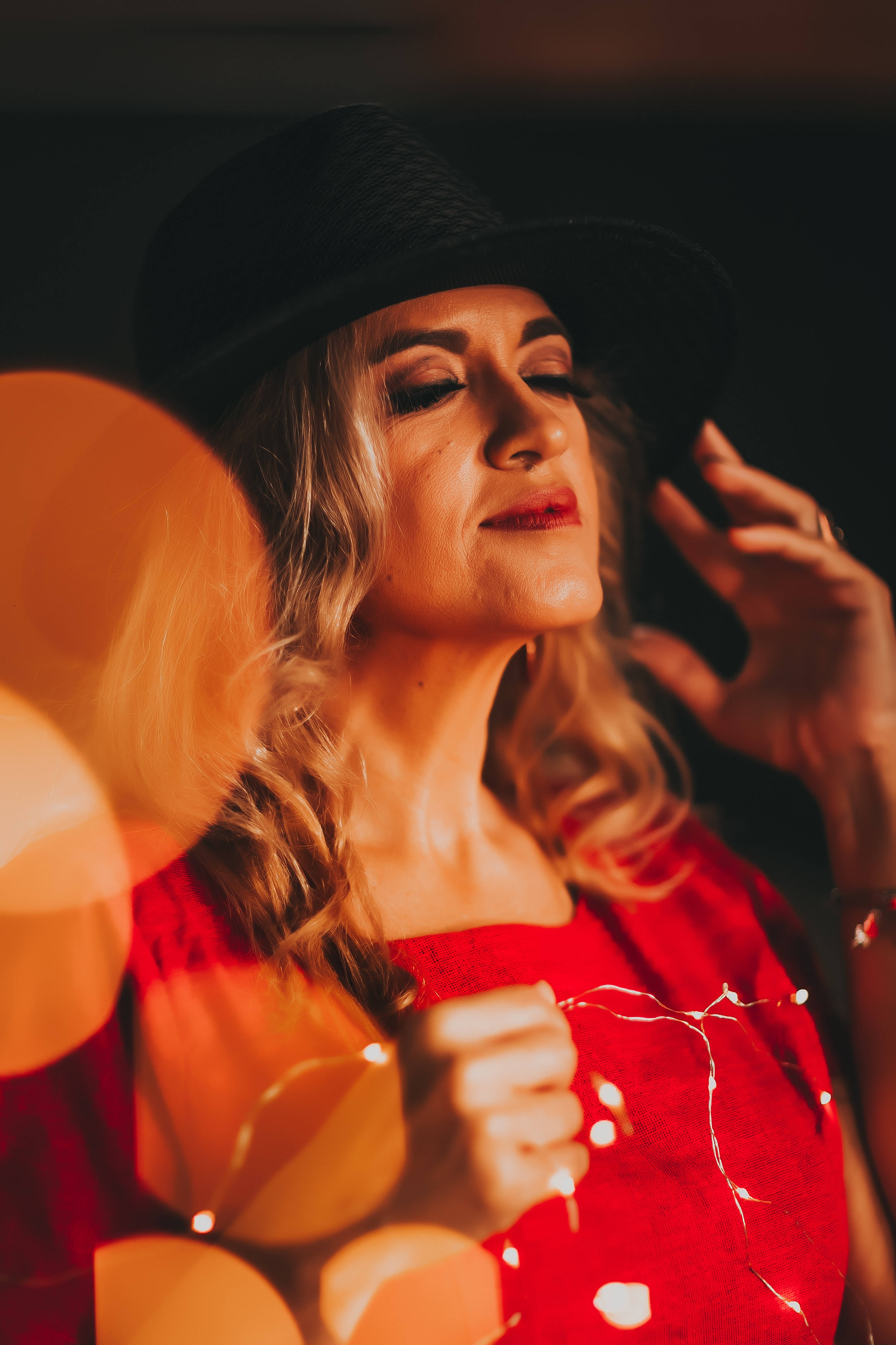 Woman 桌布 壁紙 — 一位戴黑色帽子和穿紅色連衣裙的女性，在溫暖散景的深色背景下，輕輕微笑著拿著燈串。