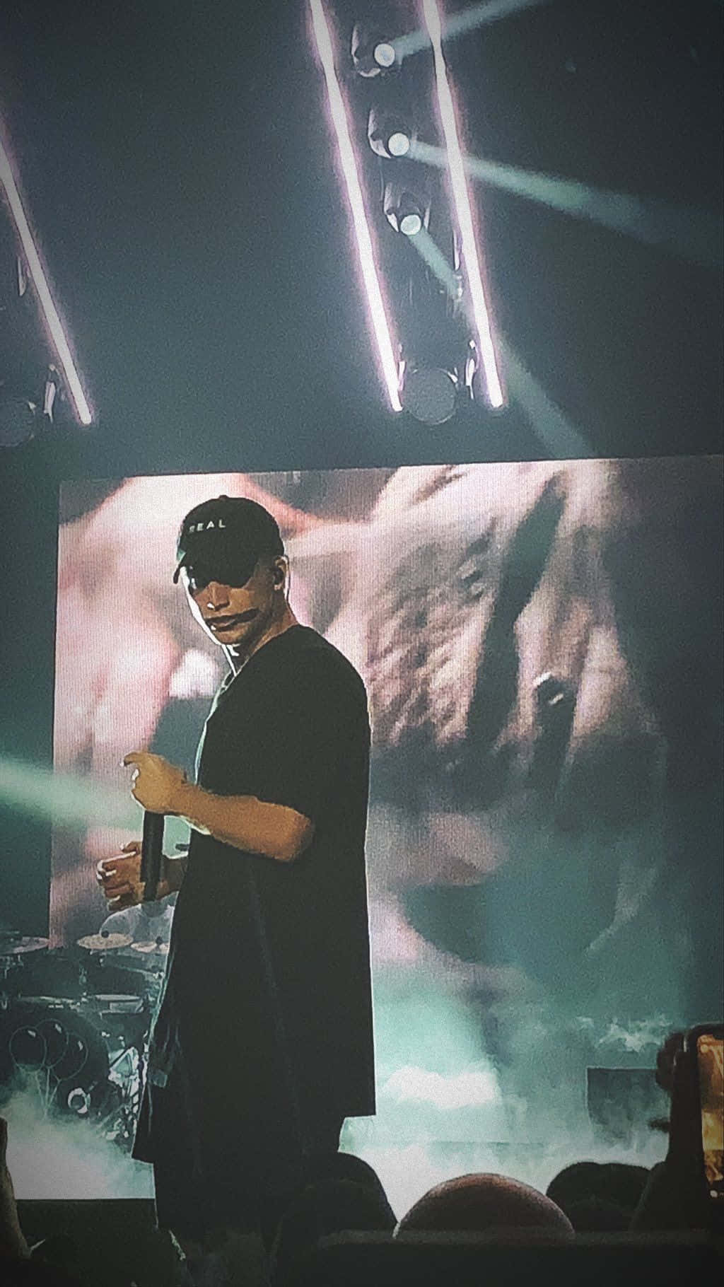 Nf Rapper 桌布 壁紙 — 一位戴黑色帽子和穿黑色服裝的表演者站在燈光昏暗的舞台上，身後有明亮的聚光燈和一個大螢幕。