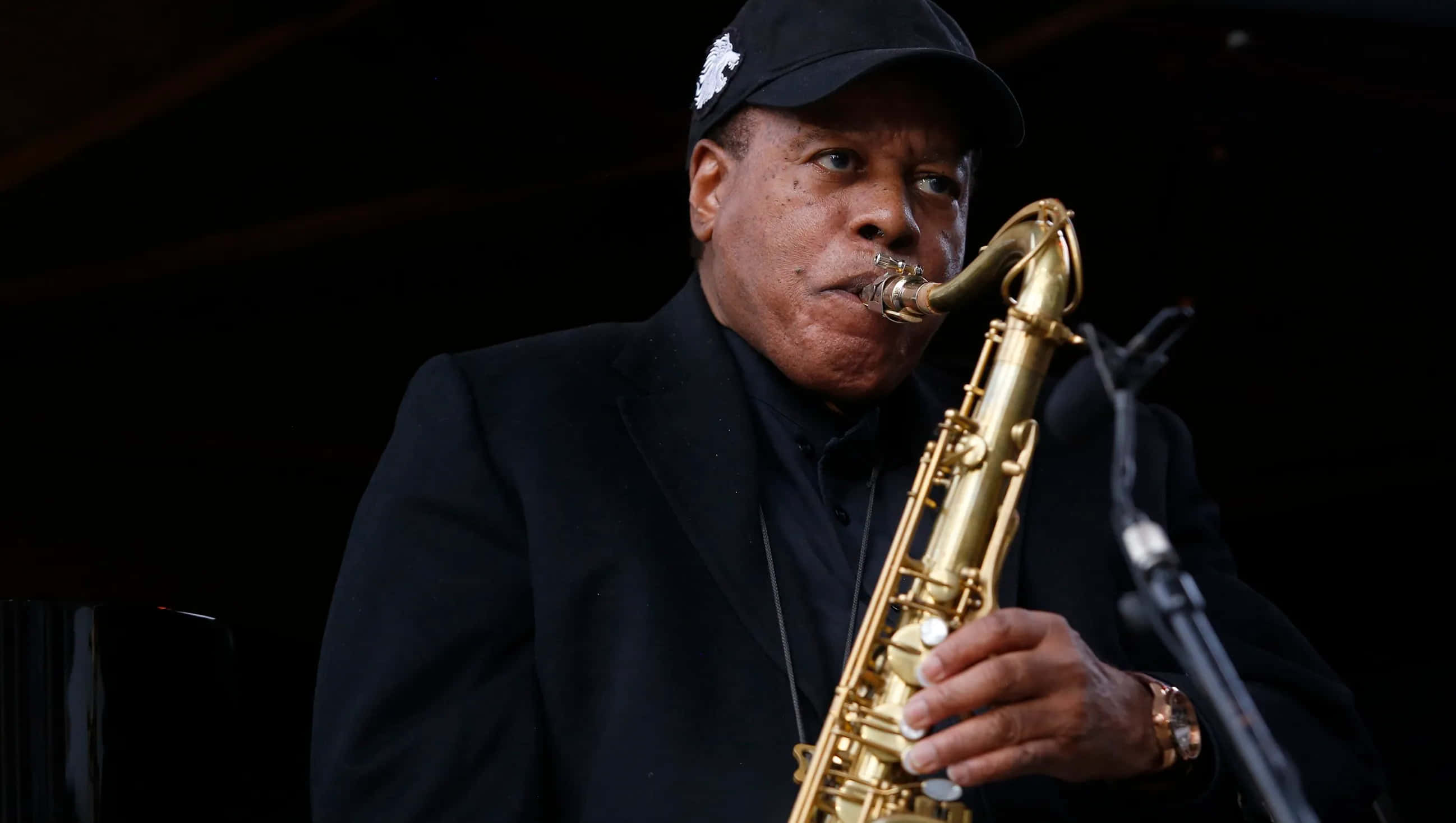 Wayne Shorter 桌布 壁紙 — 一位戴黑色帽子和西裝的爵士音樂家在舞台燈光下熱情地吹奏金色薩克斯風。