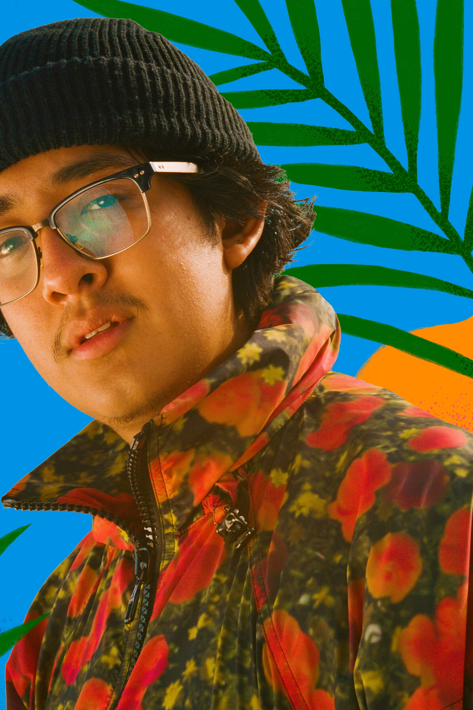Photoshoot 桌布 壁紙 — 一位戴黑色毛帽和眼鏡的年輕男子，穿著鮮豔的花卉外套，背景是帶有綠色棕櫚葉的醒目藍色。