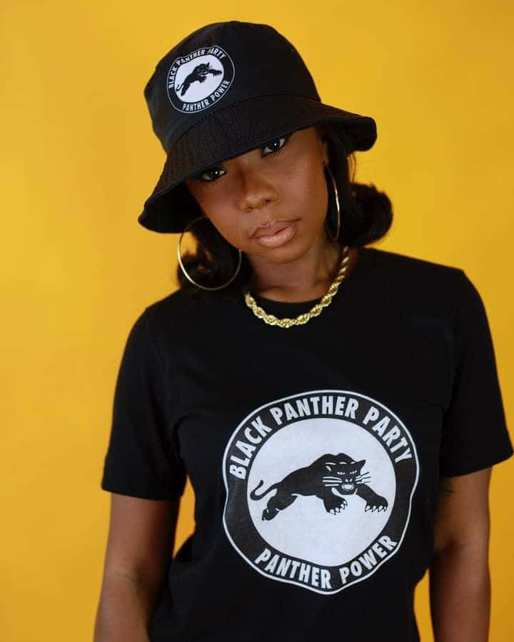 Black Panther Party 桌布 壁紙 — 一位戴黑色漁夫帽、身穿印有黑豹黨標誌T恤的女性，在鮮黃色背景前自信地站立。