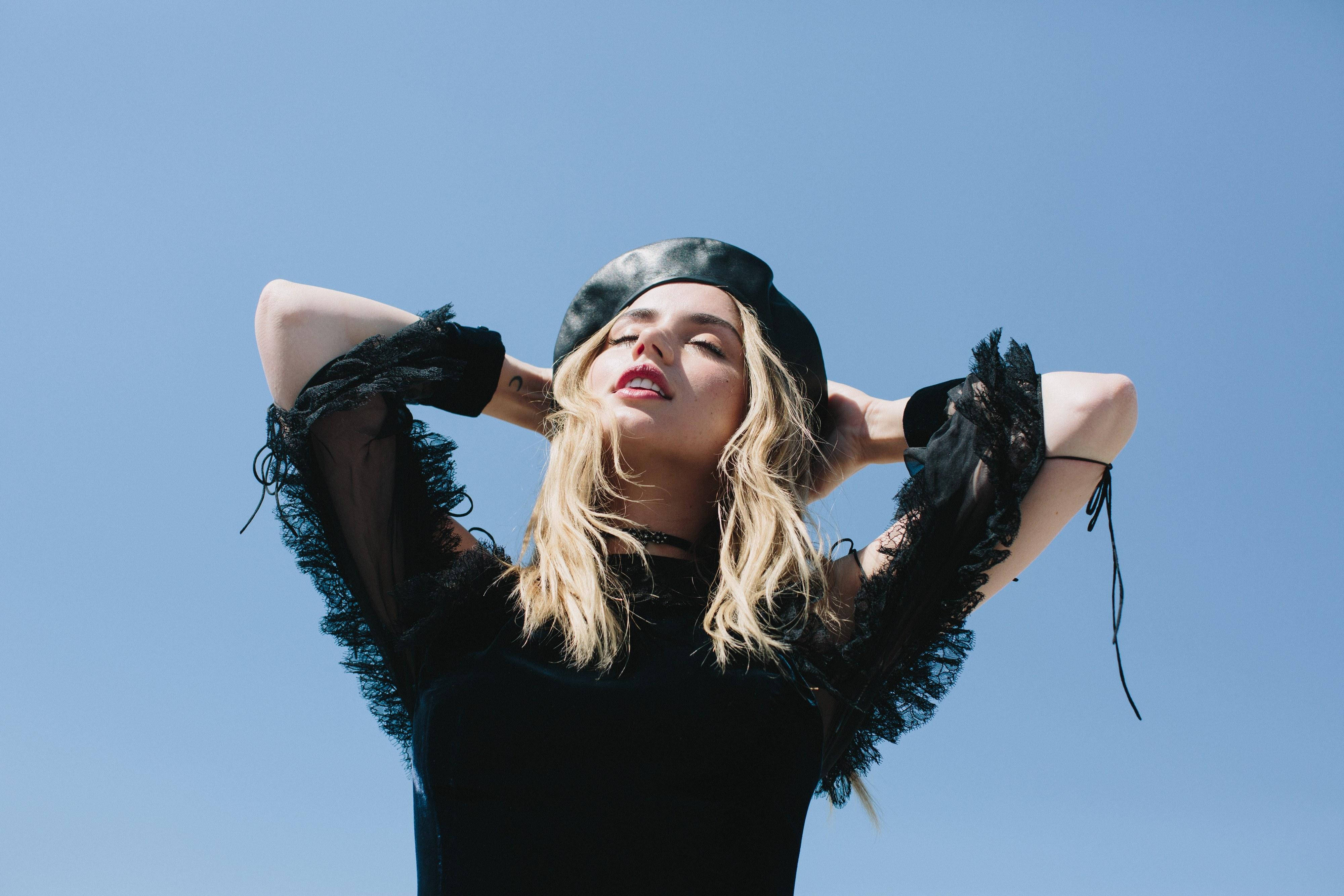 ana de armas 桌布 壁紙 — 一位戴黑色貝雷帽和蕾絲袖子的女士，閉著眼睛，雙手放在腦後，站在清澈的藍天下。