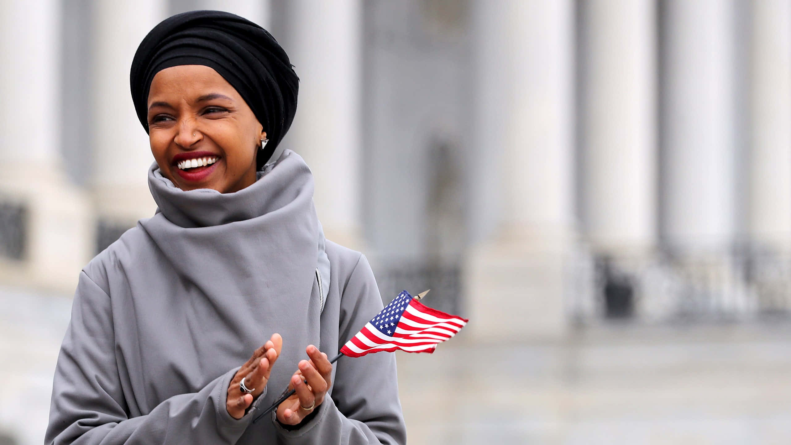 USA Flag 桌布 壁紙 — 一位戴黑色頭巾、穿灰色外套的快樂女性，在經典白色圓柱前微笑，手持一面小型美國國旗。