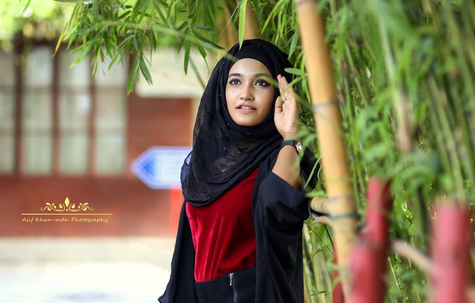 muslim 桌布 壁紙 — 一位戴黑色頭巾、身穿紅色上衣的年輕女性優雅地站在茂密的綠色竹叢中，背景是一座模糊的建築物。