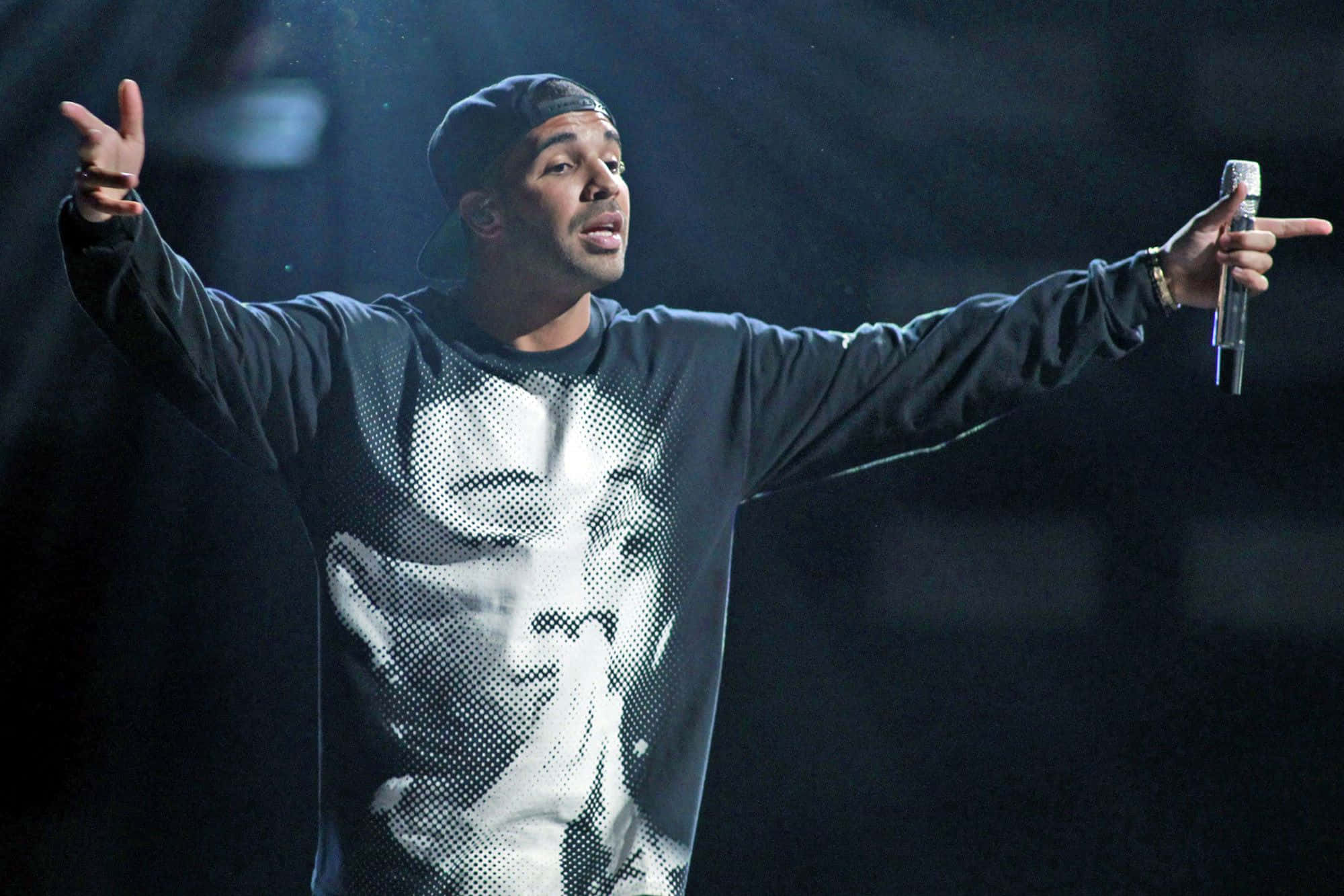Drake 桌布 壁紙 — 一位戴黑色鴨舌帽、身穿圖案連帽衫的男表演者，在黑暗的舞台上張開雙臂握著麥克風，充滿激情地演唱。