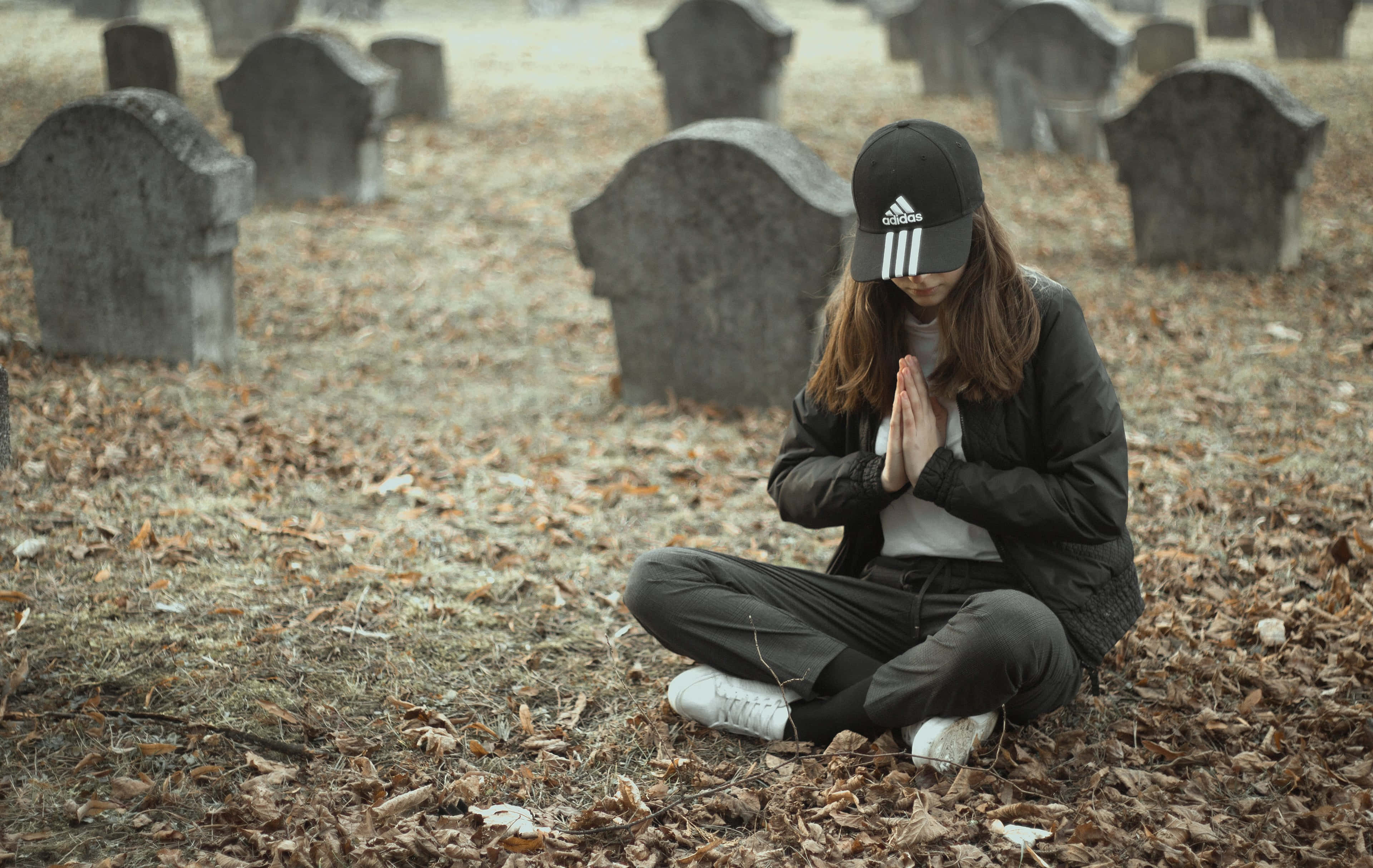 Praying 桌布 壁紙 — 一位戴黑色鴨舌帽的年輕女子在寧靜的墓園中盤腿坐在落葉間，在風化的墓碑前雙手合十祈禱。
