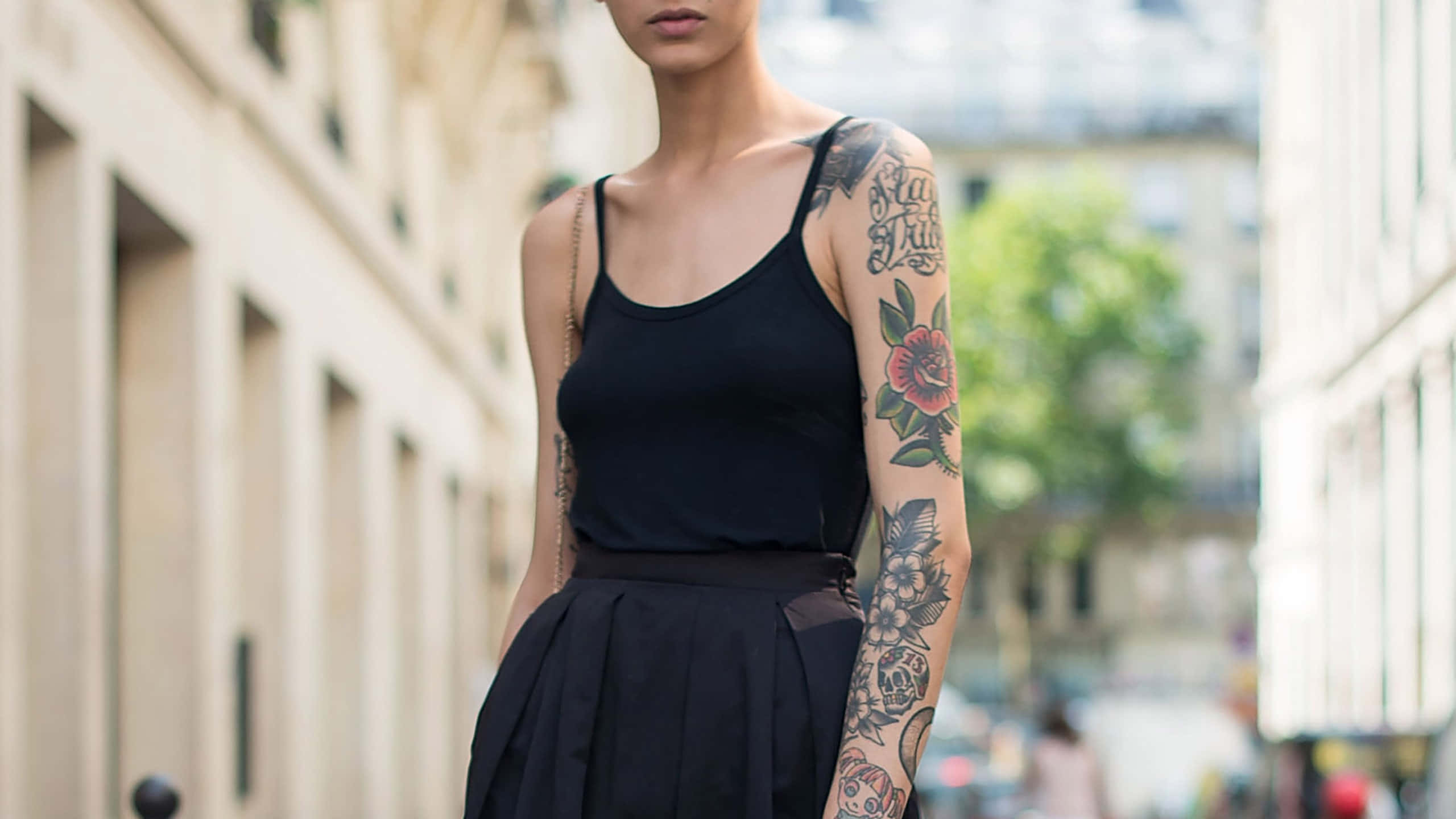 Halloween Tattoos 桌布 壁紙 — 一位手臂上有複雜刺青的女性，自信地站在陽光明媚的城市街道上，穿著黑色上衣和裙子。