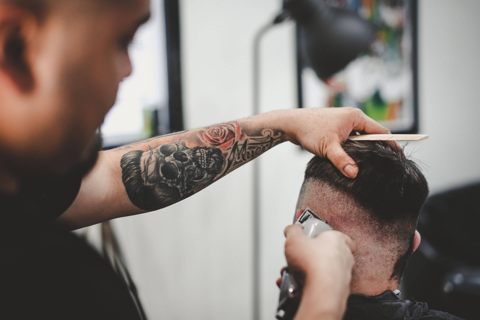 barbershop 桌布 壁紙 — 一位手臂上有骷髏頭和玫瑰刺青的理髮師，在現代理髮店用推剪為客人修剪頭髮。