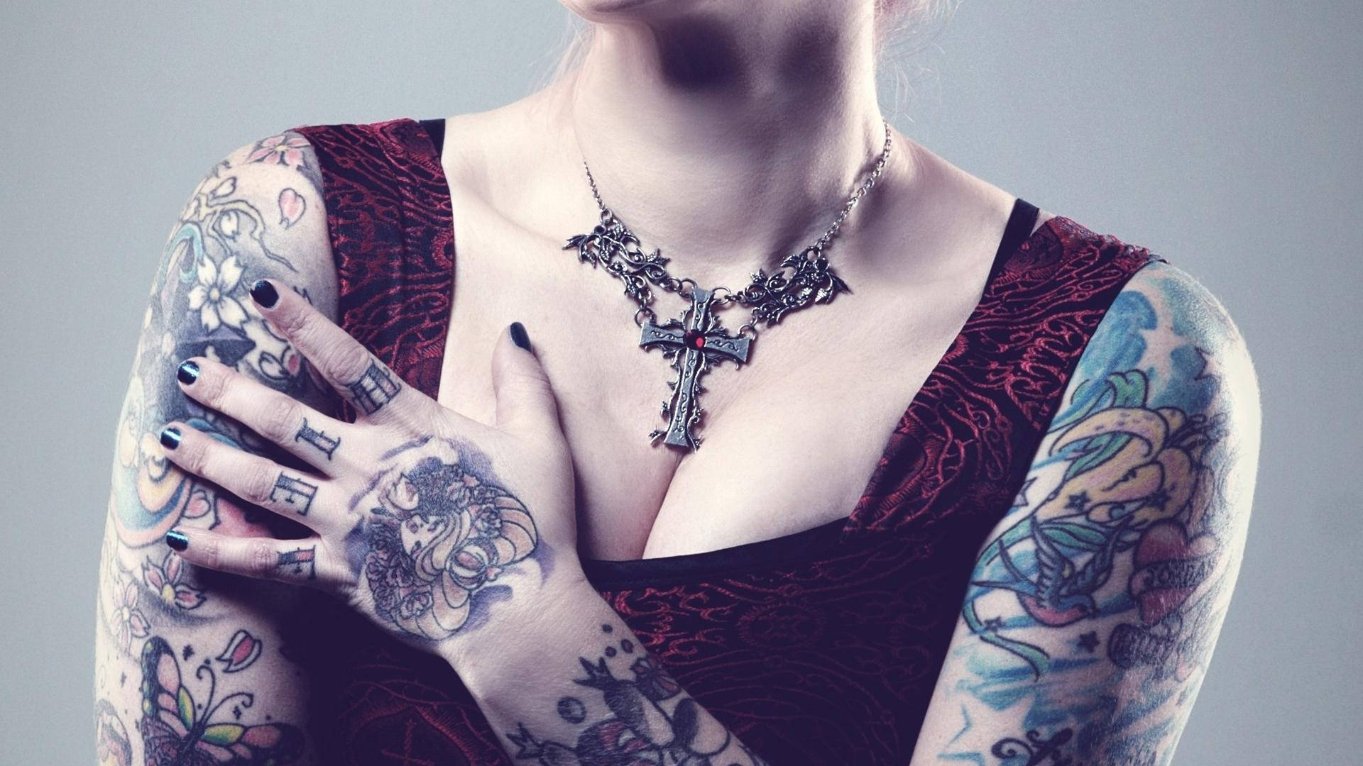 Tattoo 桌布 壁紙 — 一位手臂和手上有複雜紋身的女性，在純灰色背景前戴著哥德式十字架項鍊。
