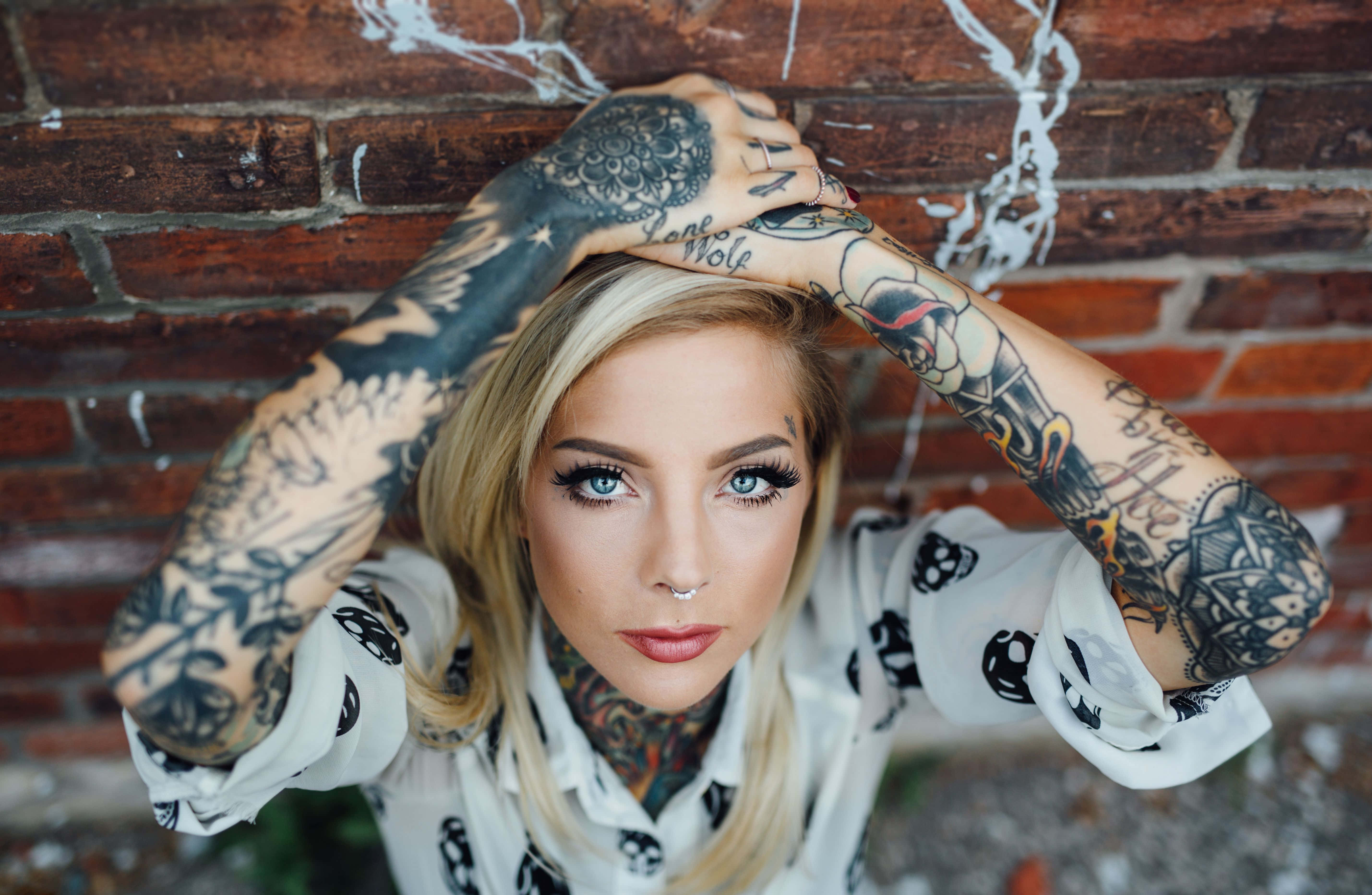 Tattoo 桌布 壁紙 — 一位手臂和頸部有大面積刺青的女性，靠在一面風化的磚牆上，直視鏡頭。