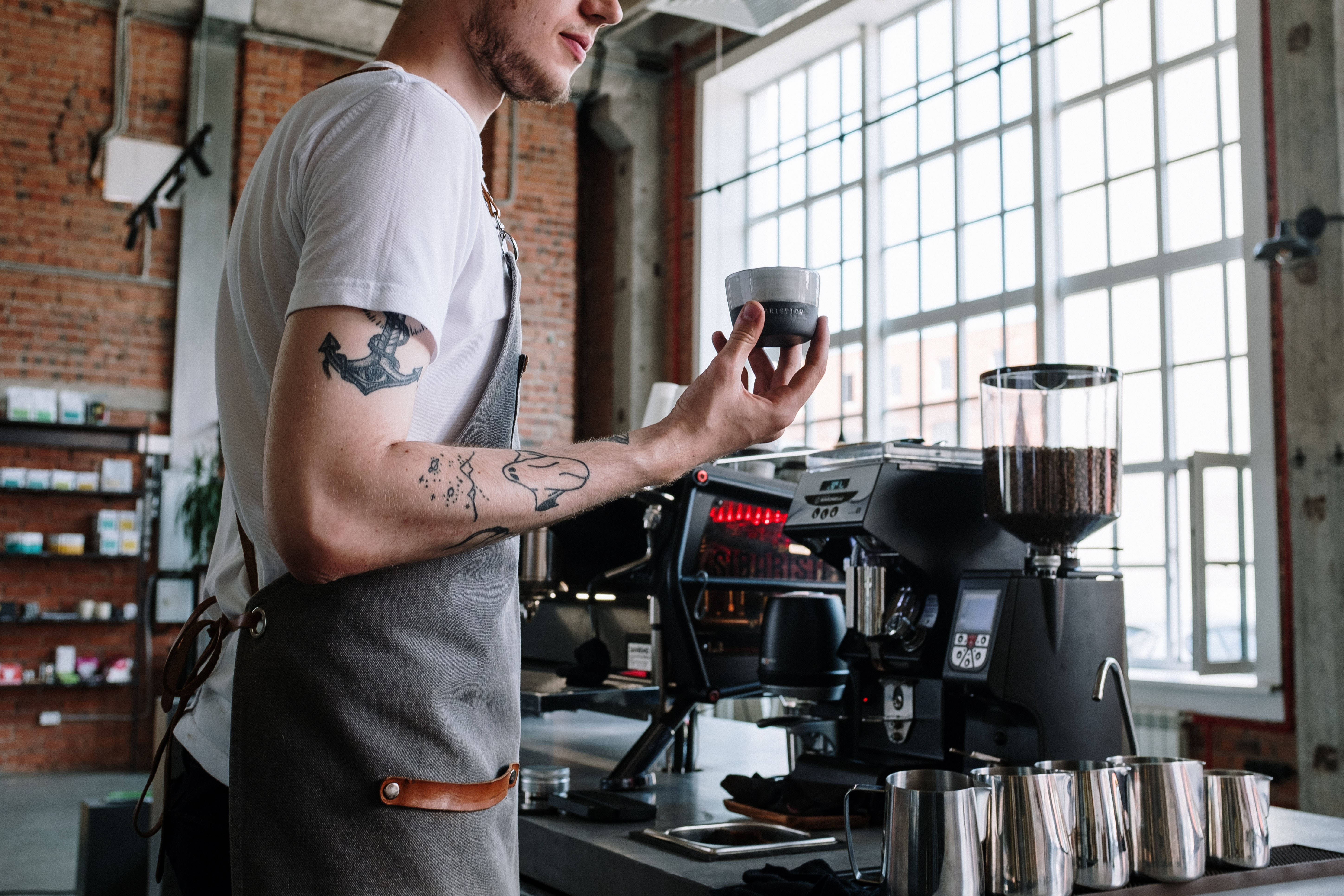 Coffee Shop 桌布 壁紙 — 一位手臂有刺青的咖啡師在明亮的工業風咖啡館中，在專業濃縮咖啡機旁邊將咖啡倒入杯中。