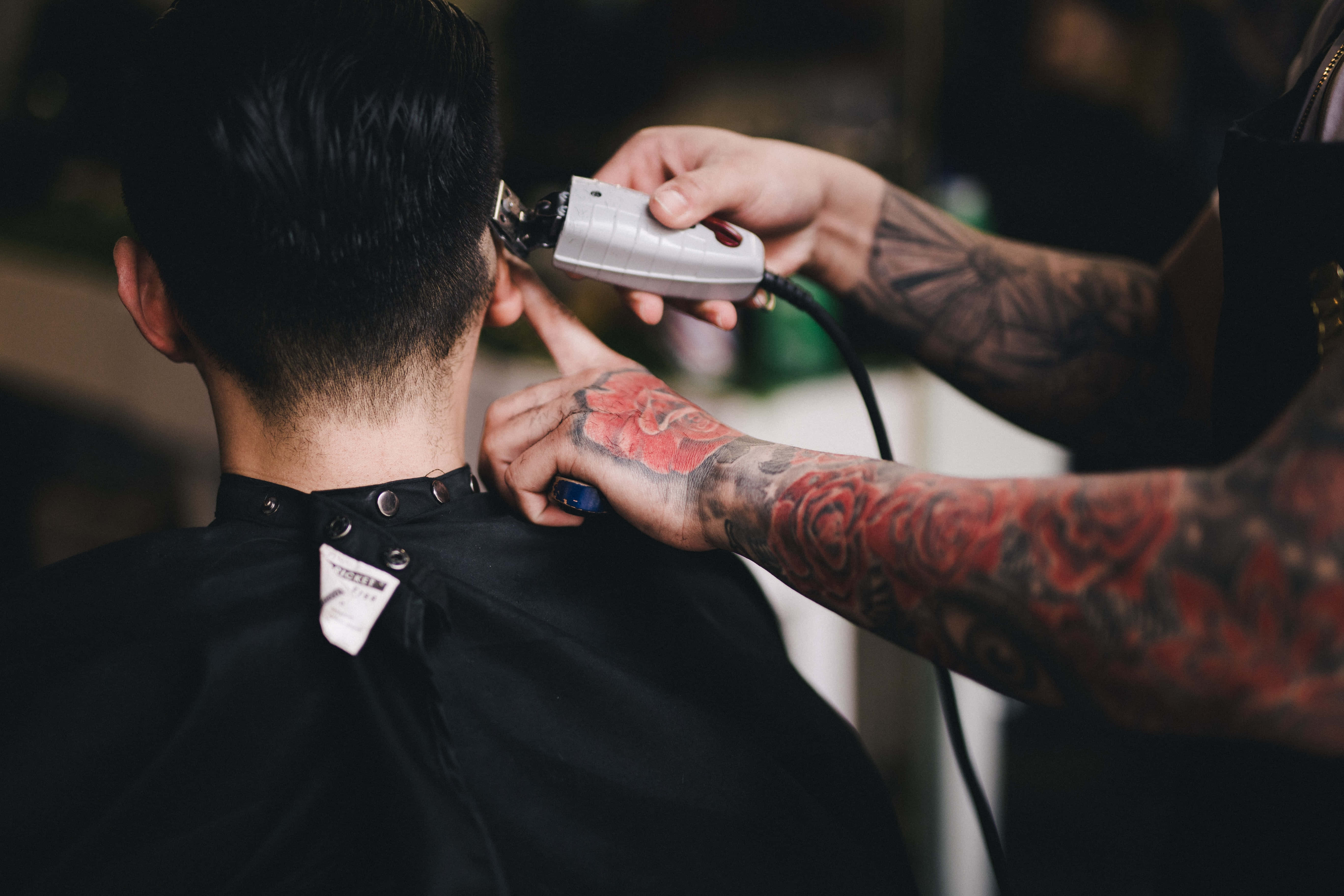 Barber 桌布 壁紙 — 一位手臂有刺青的理髮師在髮廊用電動推剪修剪顧客的短黑髮。